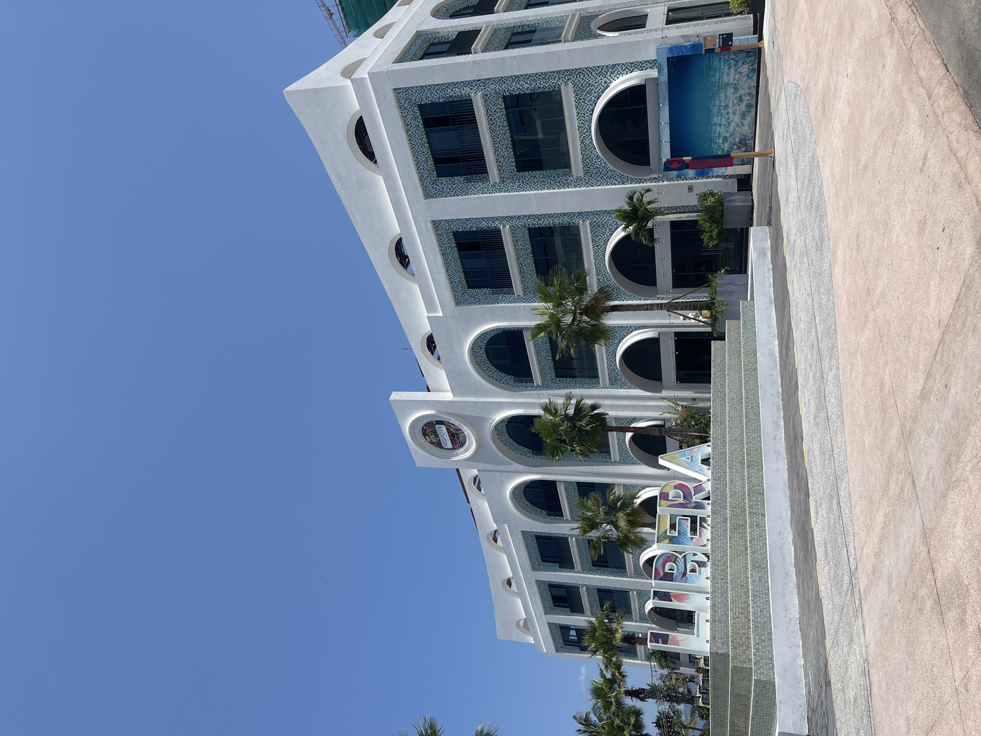 The image shows a modern, white building with blue tiled accents and arched windows, located in Nha Trang, Vietnam. In front of the building, large, colorful 3D letters spell out "LIBERA". Palm trees are visible in front of the building, and the sky is clear and blue. The area in front of the building appears to be a public space with paved walkways and decorative water features. A large mural depicting an underwater scene is mounted on the side of the building. The building appears to be a commercial or tourist establishment.