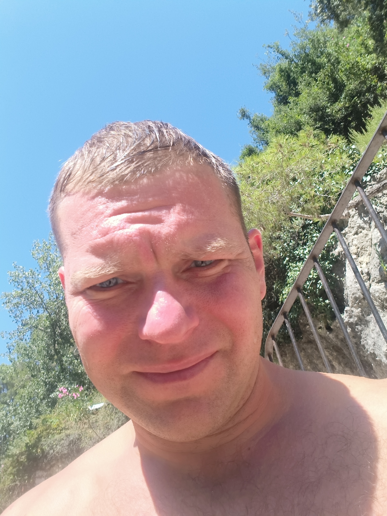 A man with short, blond hair and blue eyes is taking a selfie outdoors on a sunny day. He is shirtless and has fair skin. In the background, there are lush green trees, a rocky wall, and a metal railing. The sky is a clear blue. The scene appears to be in a natural setting, possibly a park or a hillside with vegetation. There are no other people visible in the image. The man's expression is neutral, and he is looking directly at the camera. The lighting suggests it is midday, with the sun casting shadows on his face and shoulder. The image is focused on the man's upper body and face.