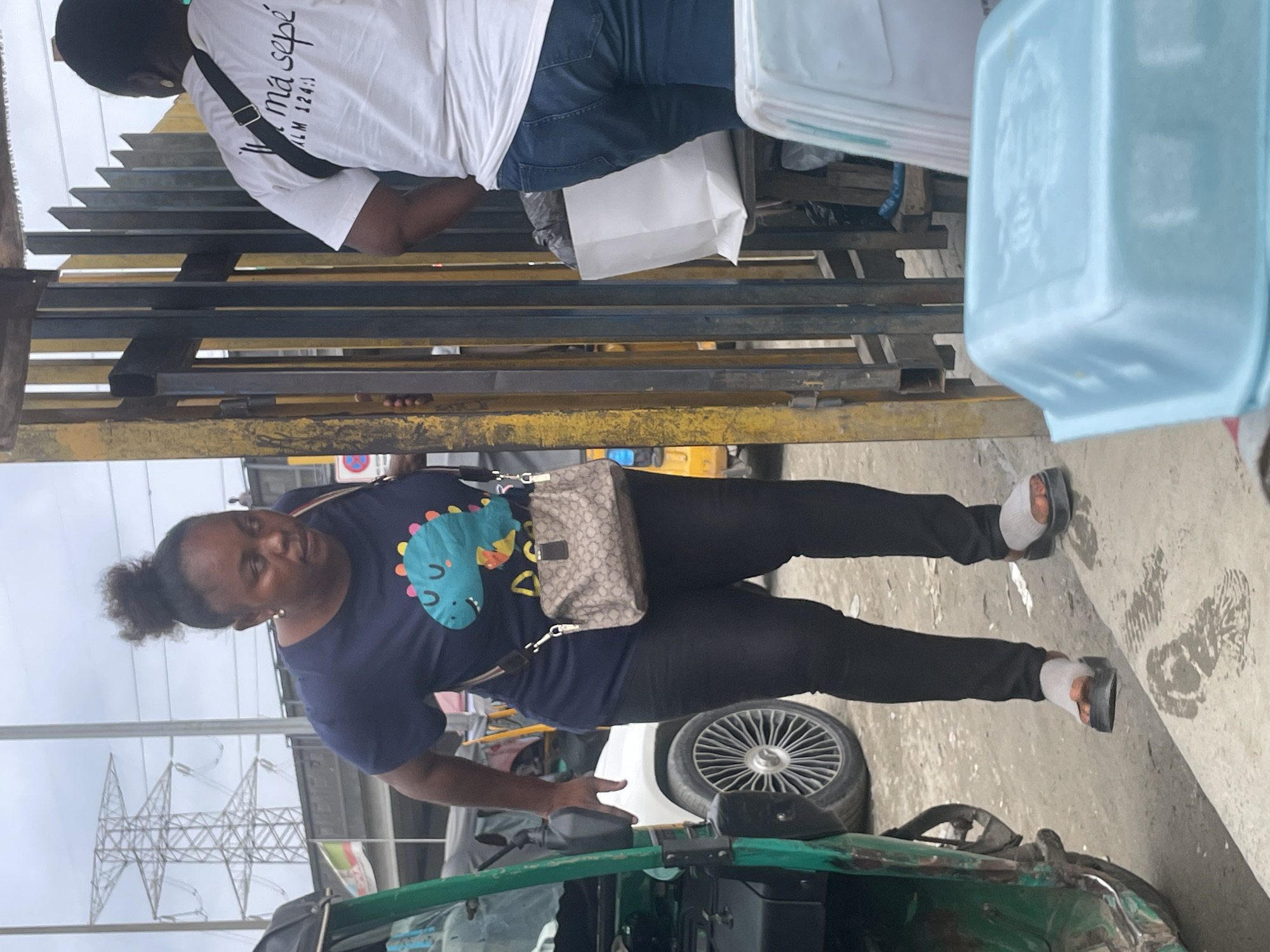 An overcast day in Ajah, Nigeria, with a woman of color in the foreground wearing a dark blue t-shirt with a light blue dinosaur graphic, black leggings, and gray and orange sandals with white socks. She is carrying a beige patterned crossbody bag. She appears to be walking past a yellow and dark gray metal gate. To her left, a green motorized rickshaw and a white vehicle are visible. In the background, a large electrical tower and the cloudy sky are present. Behind the gate, another person in a white t-shirt and blue jeans stands near a white paper bag on a stool. In the immediate foreground, a light blue plastic container is prominently displayed.