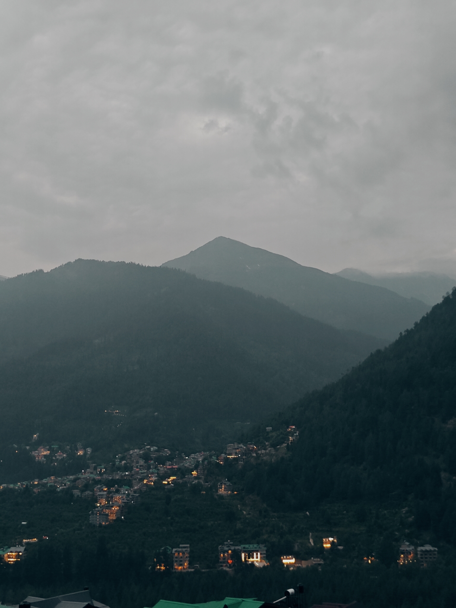 This media file captures a twilight scene of a mountain village nestled within a valley. The dominant subjects are the natural landscape and the man-made structures of the village. Towering, mist-shrouded mountains form the backdrop, their peaks fading into a cloudy, overcast sky. The slopes of the mountains are densely covered in dark green foliage, suggesting a forest.
The village itself is a collection of buildings, illuminated by warm lights, clustered on the lower slopes and valley floor. The lights create a cozy and inviting contrast against the darkening natural surroundings. The architecture appears to be varied, with some buildings seemingly modern hotels or guesthouses, characterized by multiple stories and lit windows, while other structures are smaller and more traditional. There are no visible people or specific objects of focus, the emphasis being on the overall panoramic view.
The time of day is clearly dusk or early evening, as indicated by the twilight sky and the artificial lighting of the village. The weather appears to be overcast and possibly damp, contributing to the hazy, atmospheric quality of the image. The location cues, combined with the mountainous terrain and the style of architecture, strongly suggest a settlement in a hilly or Himalayan region. The overall emotion evoked is one of tranquility and peacefulness, with a touch of mystery due to the fading light and mist. No visible text is present in the image. The context provided indicates the location as Vashisht, India.