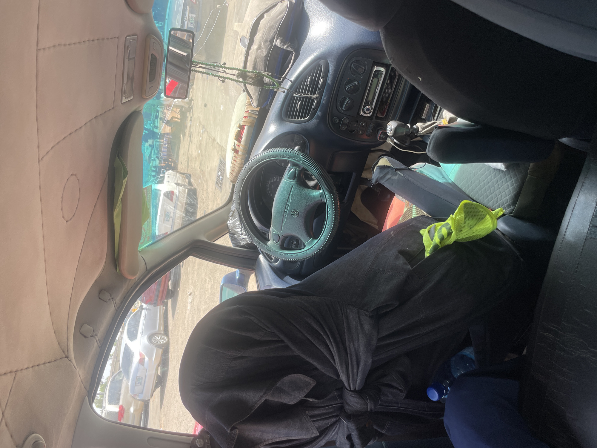 The image shows the interior of a vehicle parked in Bauchi, Nigeria. The vehicle's interior is dimly lit and appears somewhat cluttered. A person, draped in a black garment, is seated in the driver's seat, with their face obscured. The steering wheel is covered with a green, textured material. The dashboard has a prominent air vent, a car stereo, and several control knobs. A blue water bottle and a bright yellow mesh bag are visible on the floor in front of the passenger seat. Through the windshield, other vehicles and a dusty, unpaved area can be seen, suggesting the vehicle is parked outdoors, possibly in a lot or on a street. The ceiling lining of the car is visible, with stitching and some hanging items.