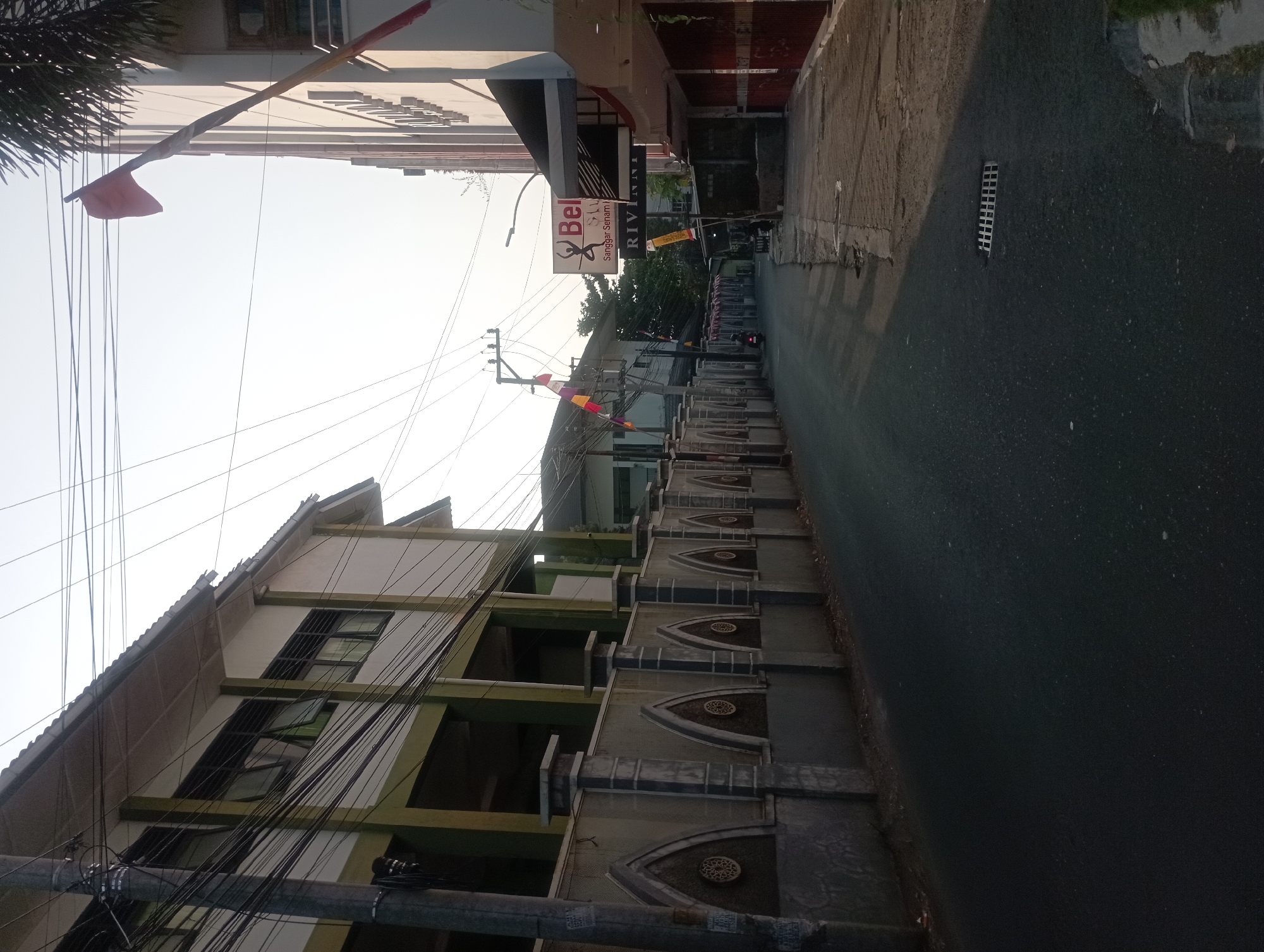 The image depicts a street scene in Yogyakarta, Indonesia, likely in a residential or commercial area, with buildings on both sides of the asphalt road. On the left, a multi-story building with green and white facade and numerous windows is visible, separated from the street by a decorative concrete wall with arched insets. Power lines and utility poles are prominent overhead. On the right, another building features a light-colored facade with several windows and awnings. A sign for "Bell" and "Sanggar Senam" (Dance Studio) is mounted on this building, along with other unreadable signs. A red flag is attached to a pole extending from this building. The street is empty except for a person on a motorcycle in the distance. The sky is bright and clear, suggesting it is daytime, possibly early morning or late afternoon due to the low angle of the sun casting shadows. The environment appears somewhat aged, with some visible wear and tear on the road and sidewalks.