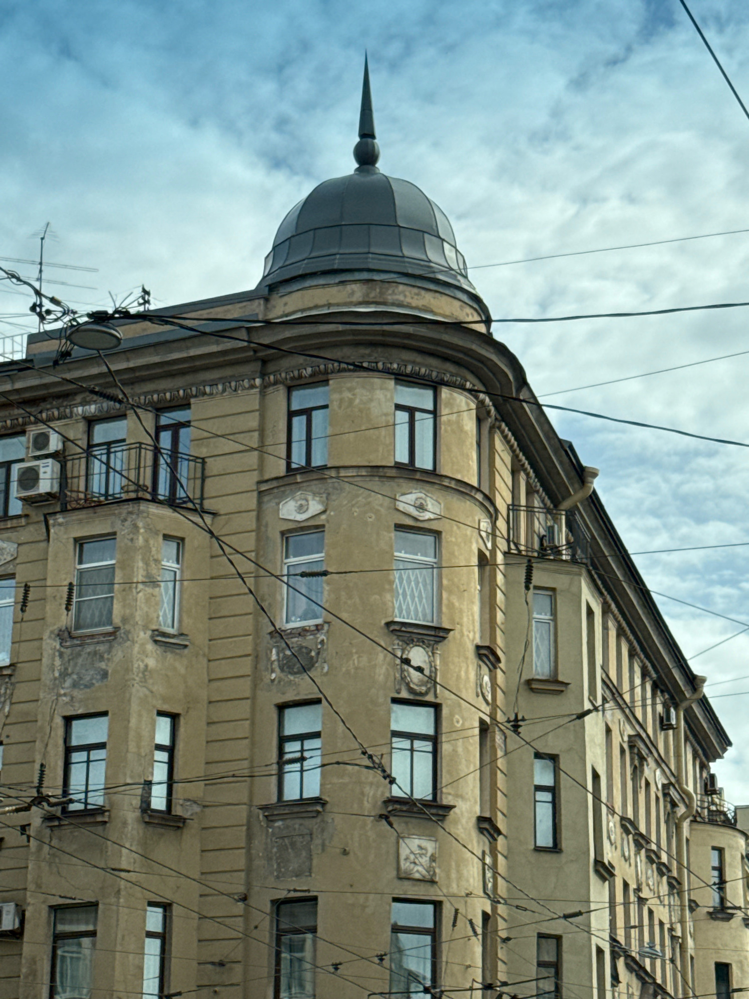 This is a low-angle, medium shot of a light brown, multi-story building with a dome and spire in Saint Petersburg, Russia. The building is weathered, with peeling paint in some areas. It features multiple windows, some with balconies, and air conditioning units are visible on one side. Numerous overhead electrical wires crisscross the frame, obscuring parts of the building and the sky. The sky is cloudy and overcast. There are no people visible in the image.