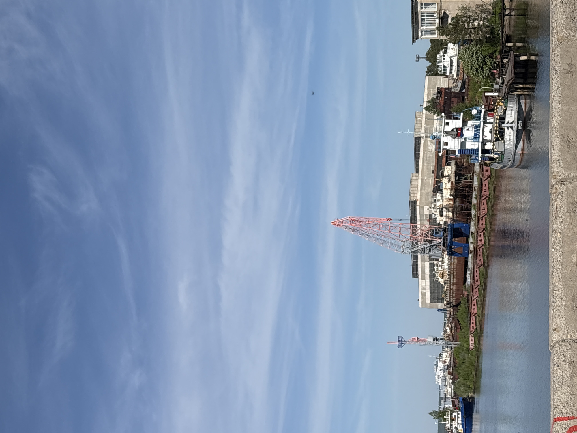 The image depicts a waterfront scene in Chaikovsky, Russia. The sky, predominantly blue, is streaked with wispy clouds. In the foreground, a concrete structure, possibly a bridge or quay, is visible. A river or canal runs parallel to the structure, reflecting the buildings and cranes on the opposite bank. A large, red lattice crane dominates the middle ground, alongside industrial buildings and warehouses. Several ships and boats are docked, indicating a functional port area. Buildings are visible, including some with white facades and windows.