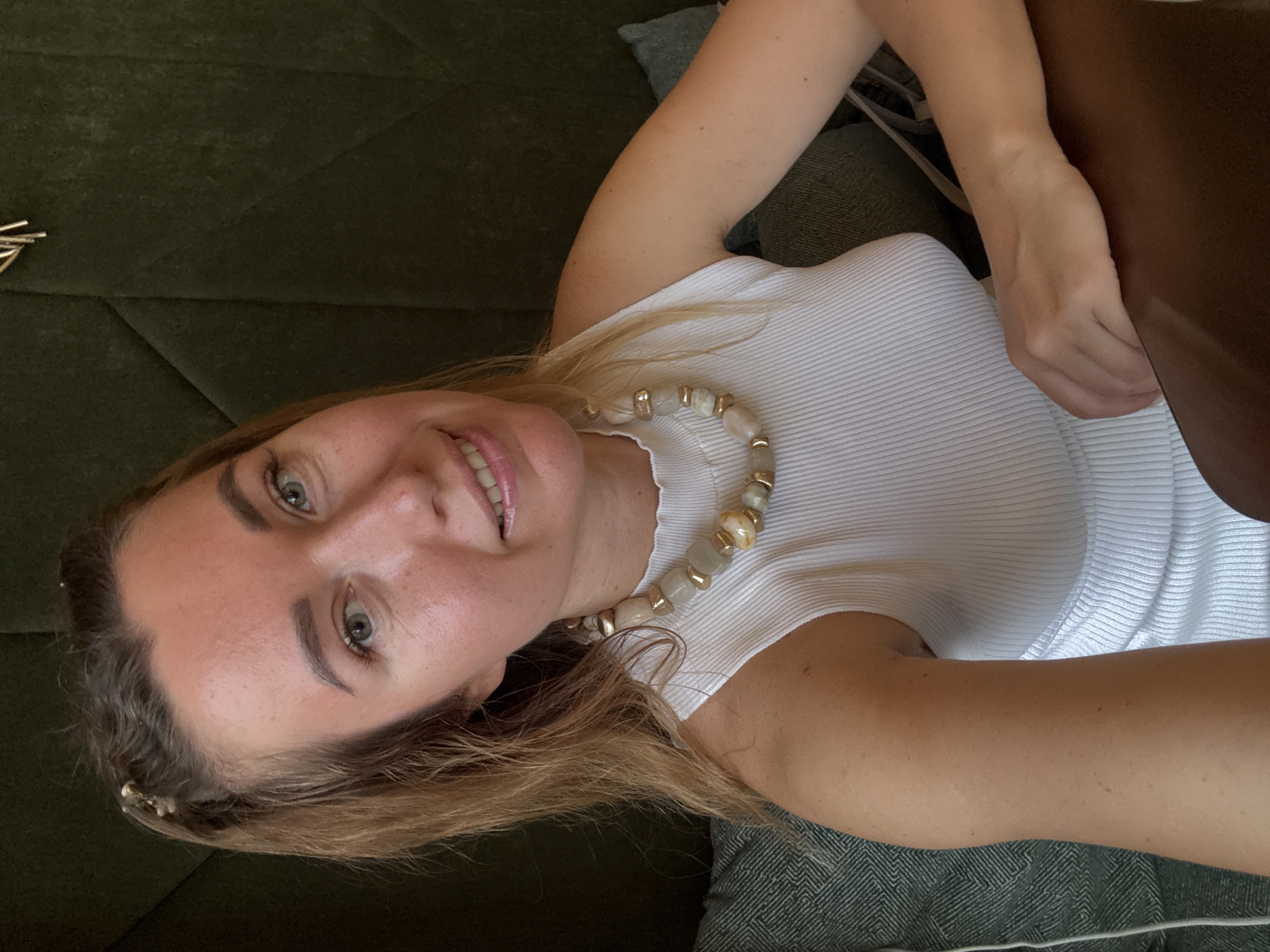 A young woman is depicted in a close-up, selfie-style photograph, taken from an overhead perspective and rotated. She lies on a dark green, textured sofa or upholstered surface.
The subject has light brown hair, styled with a few golden, pin-like accessories near her forehead on the left side of the frame. She has light-colored eyes, possibly green or hazel, and her face shows a relaxed, gentle smile with visible teeth. Her lips are a natural pink shade, and her complexion appears clear. She is wearing a fitted, white ribbed tank top or camisole. Around her neck is a distinctive necklace made of large, irregular, light-colored (possibly white or pale amber) beads, interspersed with smaller gold-colored accents.
Her right arm is visible, resting on the sofa, and her left arm is bent, with her hand partially obscured by a dark brown object on the right side of the image, which could be part of a bag or furniture. The lighting suggests an indoor setting during daylight hours, casting a soft glow on her face. The overall mood conveyed is pleasant and relaxed. There is no visible text in the image.