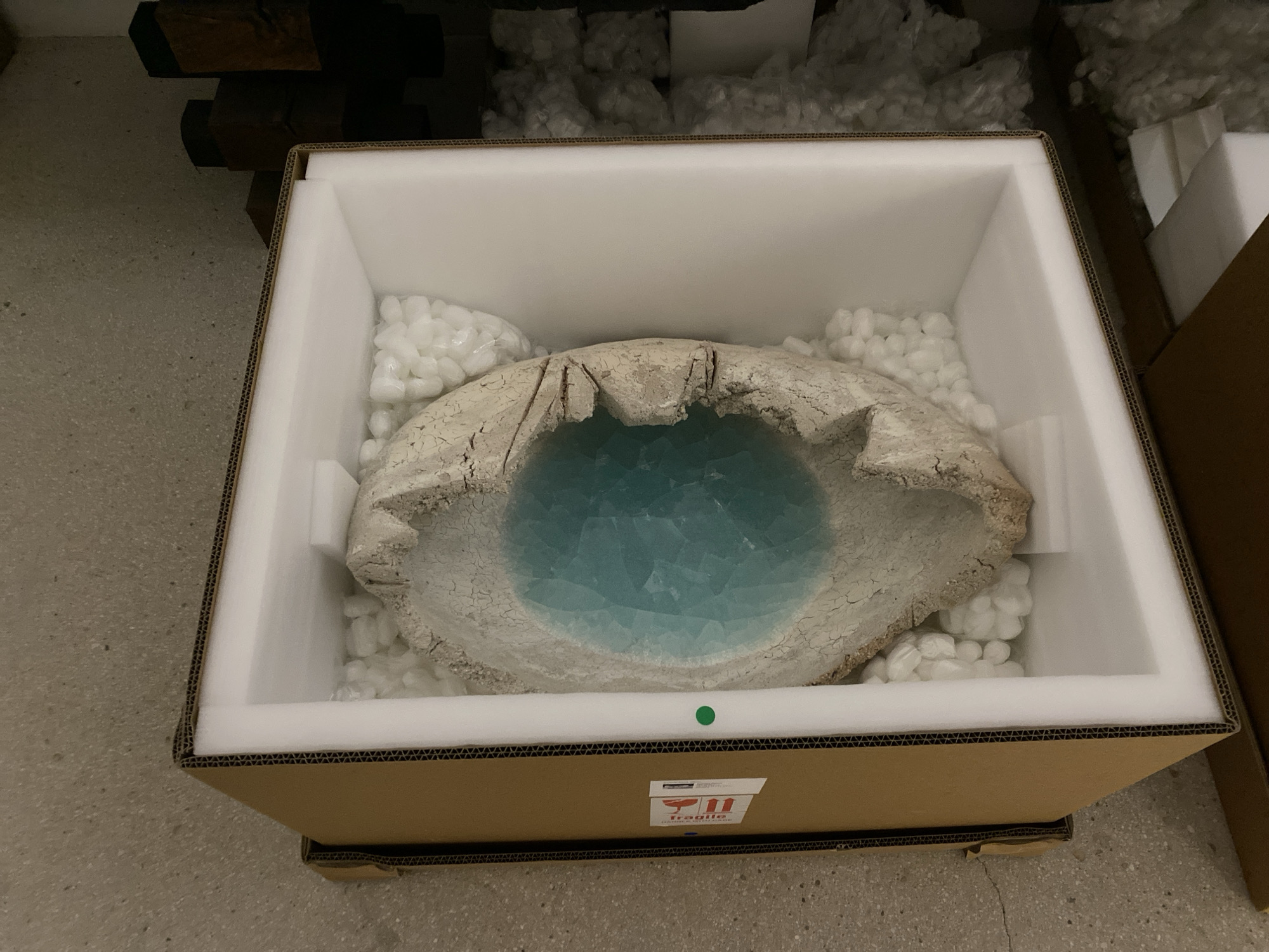 An eye-level indoor shot displays a unique, fragile art object carefully packaged within a cardboard box, likely in a storage area or workshop in Paris, France. The central object is a large, irregularly shaped piece resembling an organic bowl or basin. Its exterior is a textured, off-white or beige, with a dry, crackled appearance, while the interior is a striking, deeply crackled turquoise blue, reminiscent of ice or water.
This intricate item is securely nested within custom-cut white foam inserts inside a standard brown cardboard box. Additional white packing peanuts surround the object, providing extra cushioning. On the front of the cardboard box, a prominent red and white "FRAGILE" label is visible, featuring an upward-pointing arrow and text that hints at careful handling instructions. A small green circular sticker is also affixed to the box.
The background is dimly lit and somewhat blurred, showing what appears to be stacked dark wooden beams or similar structural elements, along with other storage materials or boxes. The floor is a light gray, possibly concrete or industrial tiling. No individuals are present in the image, and the artificial lighting suggests an indoor setting. The scene emphasizes the careful preparation and transport of a delicate and artistic creation.