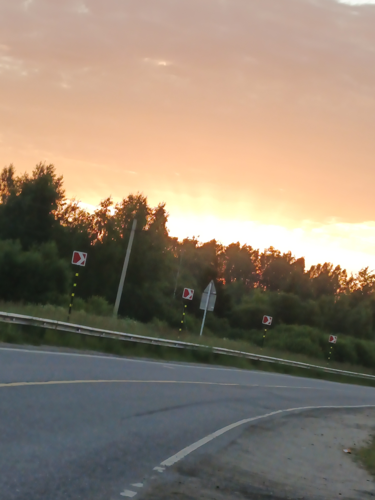 The image shows a road at sunset, with a gradient of orange and pink in the sky. The road curves to the right, with a yellow line marking the center. On the left side of the road, there is a guardrail and a line of trees and bushes. Several road signs, indicating a curve to the right, are visible along the side of the road, each accompanied by a yellow marker. There are no people or animals in the image. The overall scene depicts a rural or semi-rural road during twilight.