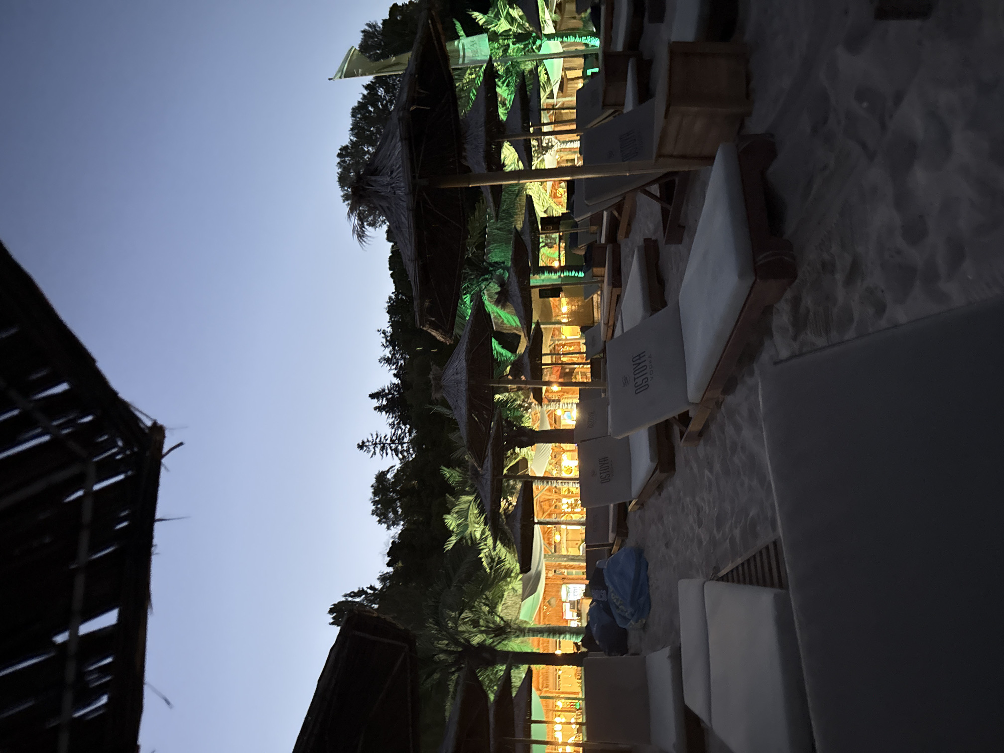 A beach resort at dusk in Varna, Bulgaria, is depicted. Rows of empty lounge chairs with white cushions are set up on the sand, some featuring the "OSTOYA" logo. Thatch-roofed umbrellas are positioned over some of the chairs. In the background, illuminated structures, possibly a bar or restaurant, glow with warm yellow and green lights, casting an ambiance over the palm trees and lush foliage. A few blue beanbag chairs are visible near the middle of the frame. The sky is a deep twilight blue.