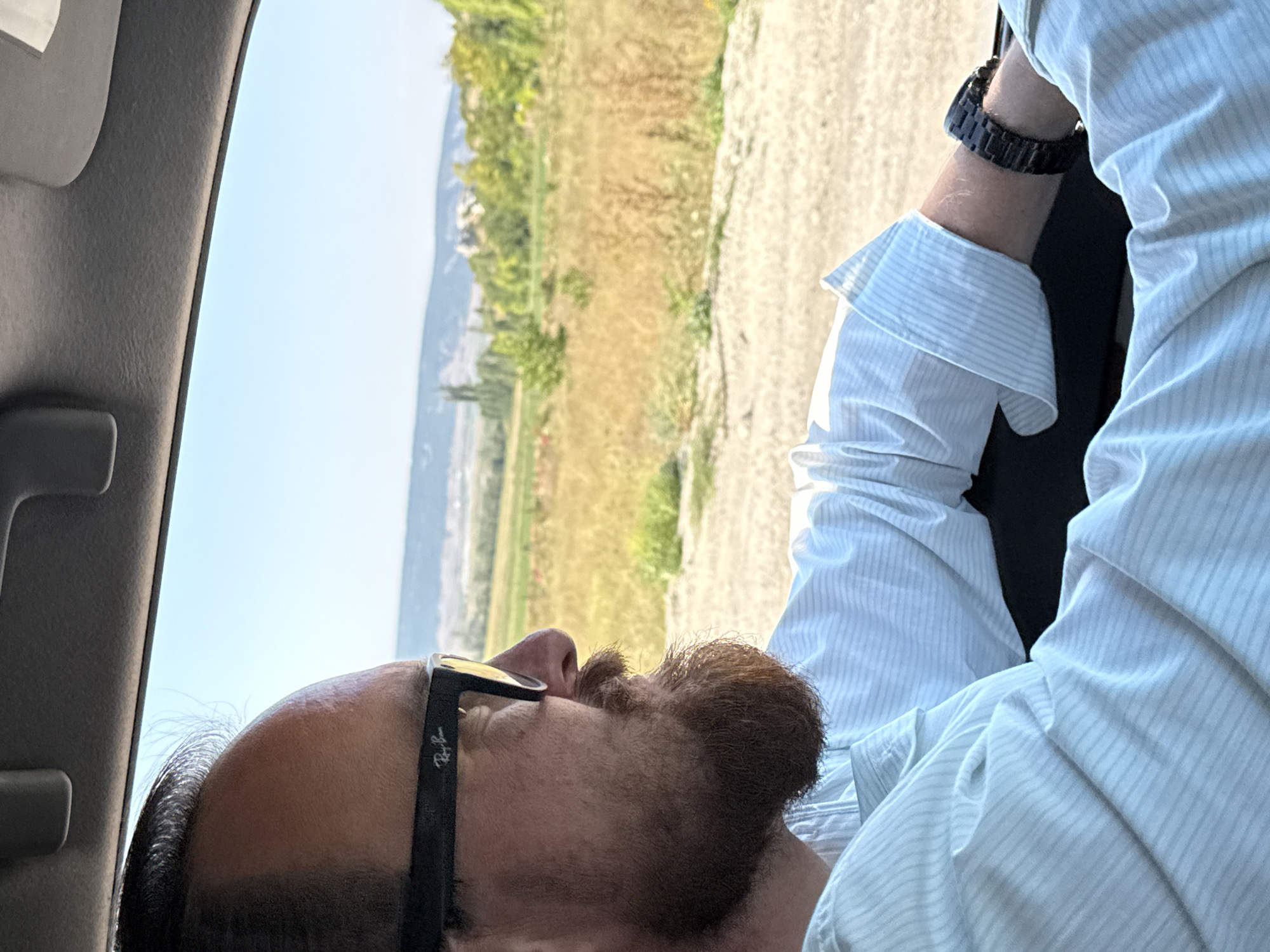 The image shows a man with a beard, wearing glasses and a light blue striped shirt, driving a car. The interior of the car is visible, with the dashboard and roof liner in the foreground. The man is looking forward, and his right arm is resting on the steering wheel. The background shows a rural landscape with fields, trees, and hills under a clear blue sky. The image appears to have been taken during the daytime. Based on the context of Красномакское сельское поселение, Ukraine, this scene likely depicts a person driving through the countryside in that region.