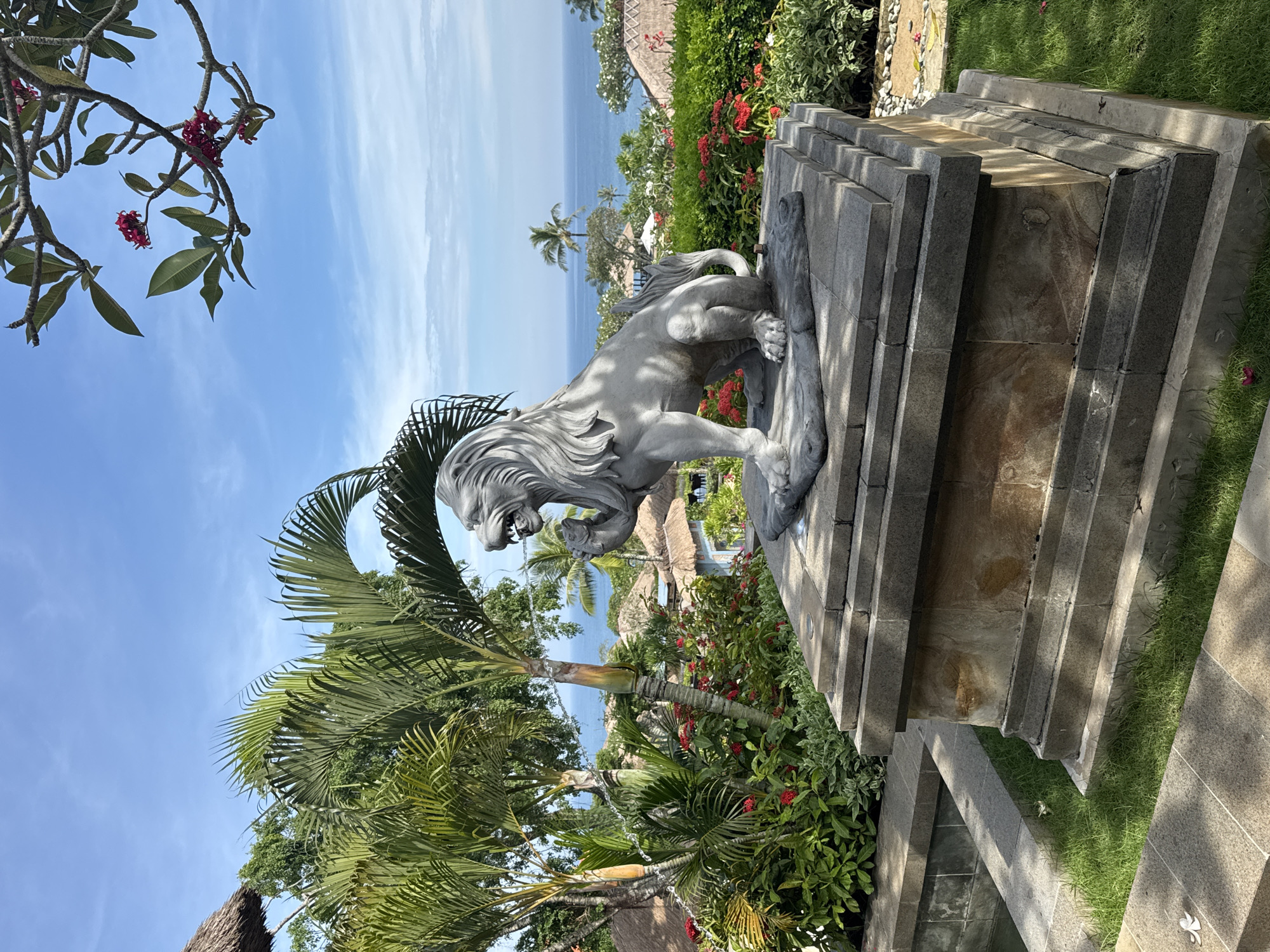 A full shot of a stone lion statue located in Kuta Selatan, Indonesia. The lion is on top of a tiered stone platform and has a menacing expression. A palm tree is to the left of the lion statue, and the ocean is in the background. Red flowers and green plants are surrounding the statue. The sky is blue with a few clouds.