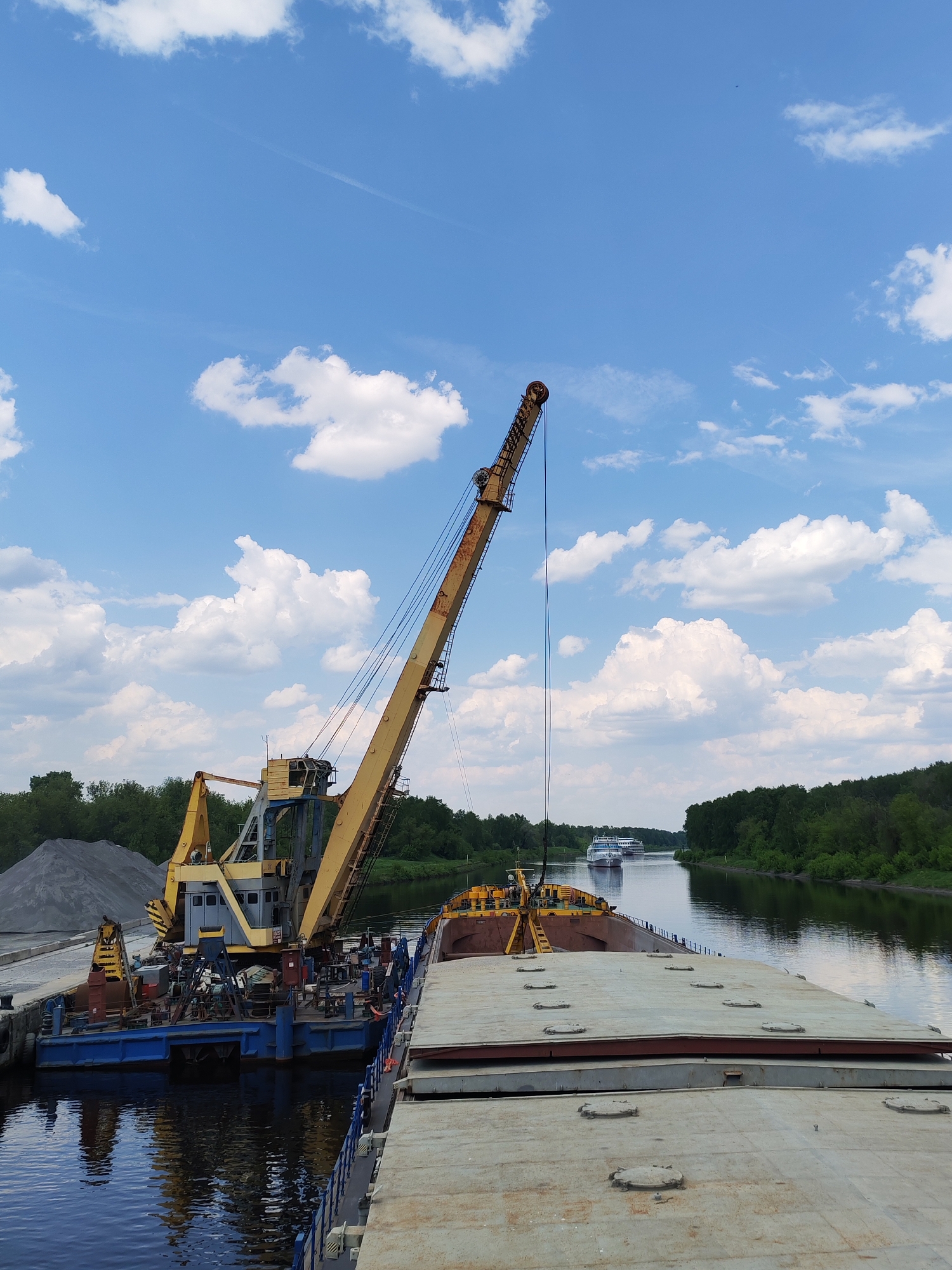 The image depicts a waterway scene near Yakhroma, Russia. A yellow and gray crane is stationed on a blue barge next to a larger, flat-topped barge. The crane is positioned near a pile of gray aggregate. In the background, a calm river flows between green, tree-lined banks, with two small passenger ships navigating the waterway. The sky is blue with scattered white clouds.