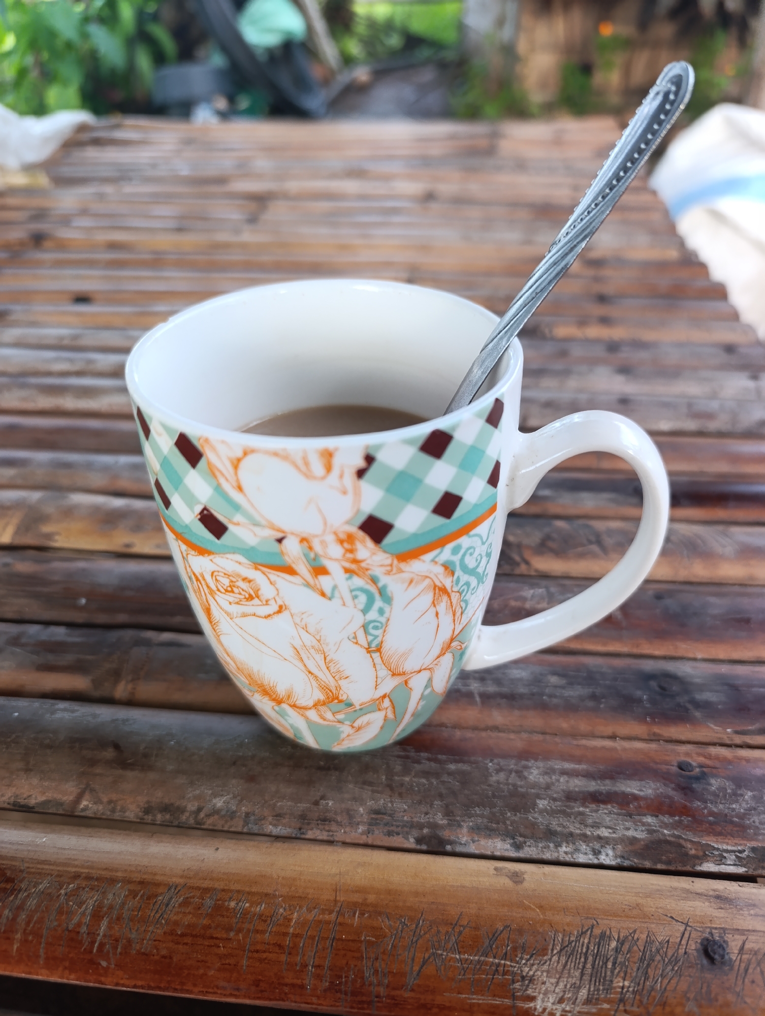 An overhead medium shot captures a close-up of a white ceramic mug with a decorative pattern, filled with a light brown liquid, likely coffee or tea. A silver spoon rests inside the mug, its handle angled towards the upper right of the frame. The mug is placed on a surface made of horizontally arranged bamboo poles, appearing to be a table or bench. The background is blurred, showing hints of greenery and possibly some outdoor structures. The lighting suggests it is daytime.