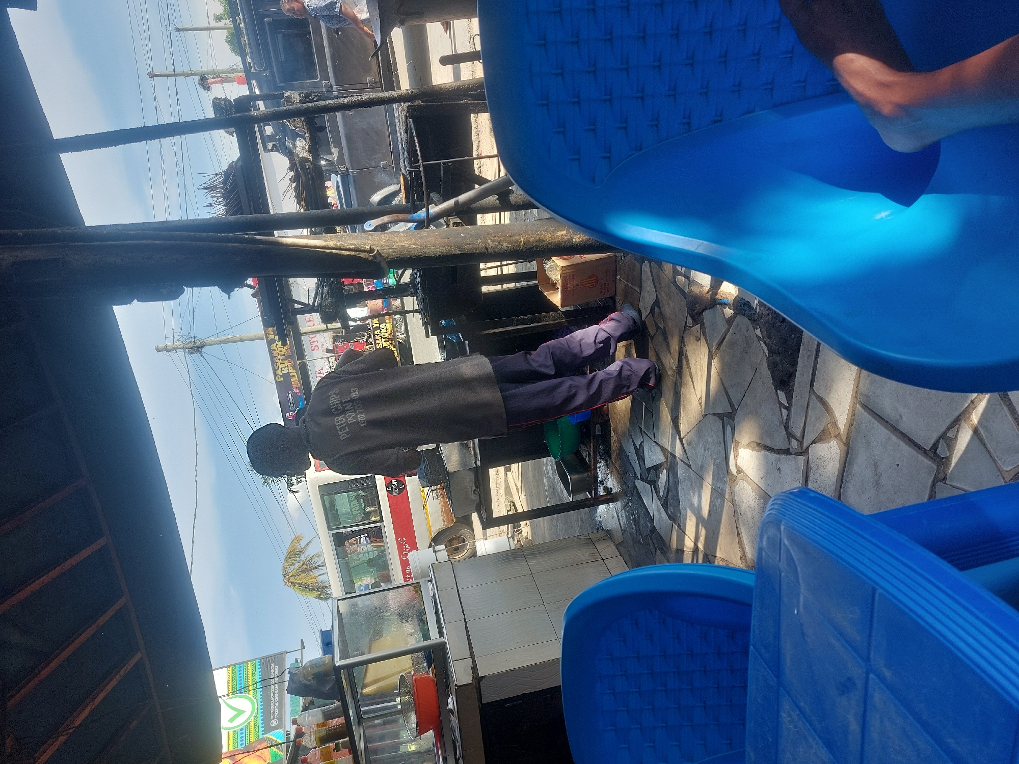 The scene appears to be an outdoor setting, possibly a roadside or street-side location in Dar es-Salaam, Tanzania. The image contains a person wearing a dark jacket and pants, standing near what seems to be a food stall or shop. There are blue plastic chairs in the foreground. A white and red bus is visible in the background, along with signage displaying the text "PETER CHIPS POINT 0712703 083". The area is paved with stone tiles. The presence of food items in a display case suggests a commercial establishment.