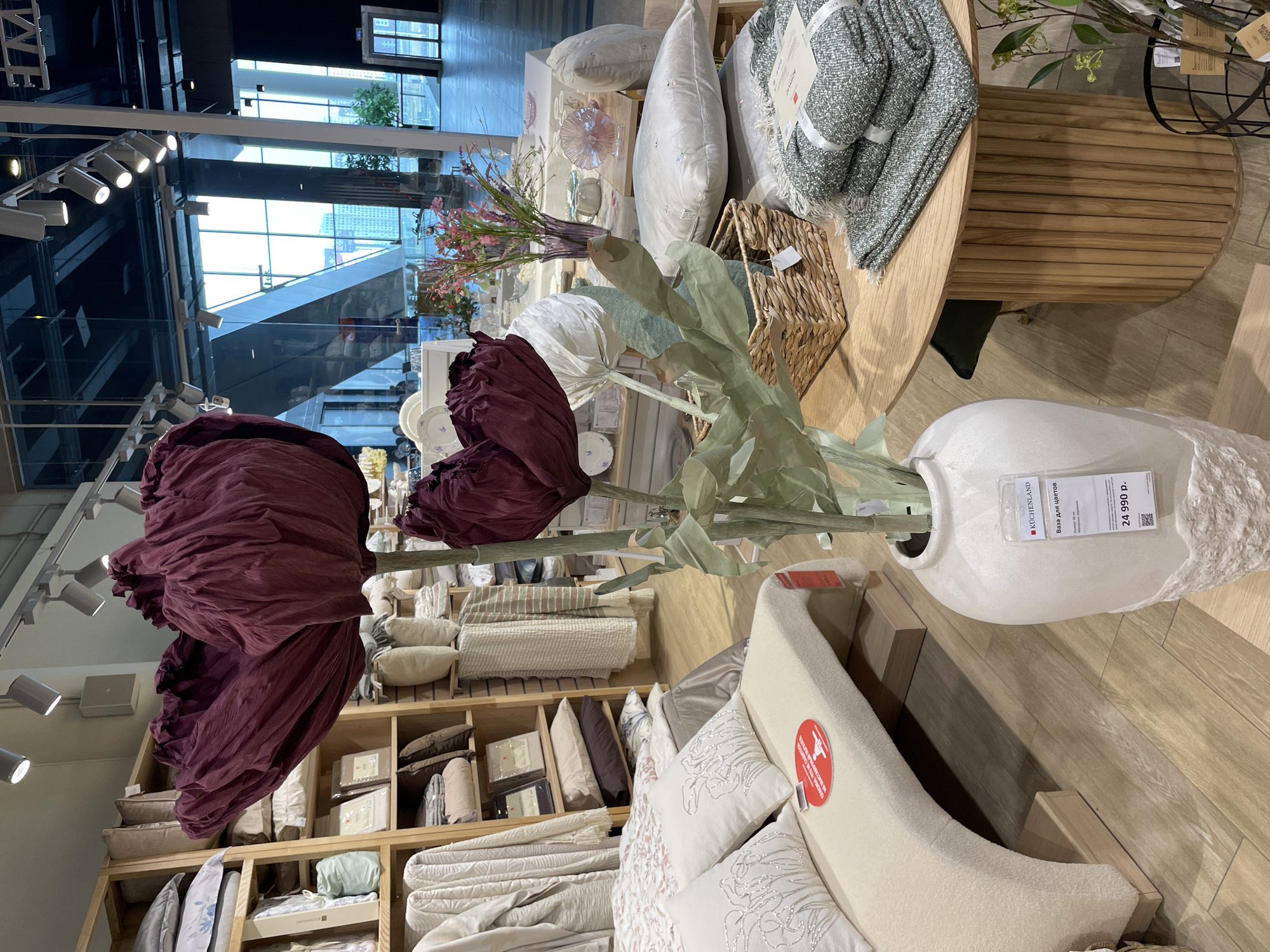 The image depicts an indoor scene, likely a home goods store in Kommunarka, Russia. The scene features a variety of merchandise displayed for sale. The focal point is a large floral arrangement with deep purple fabric flowers in a white textured vase, with signage indicating a price of 24,990 rubles. The vase is positioned on a wooden floor near a bed frame and decorative pillows. In the background, there are shelves filled with bedding, cushions, and other home decor items. A round wooden table showcases blankets, pillows, and a woven basket. A large window provides a view of buildings and a skyway, suggesting an urban setting. The overall impression is that of a retail space designed to present a curated selection of home furnishings.