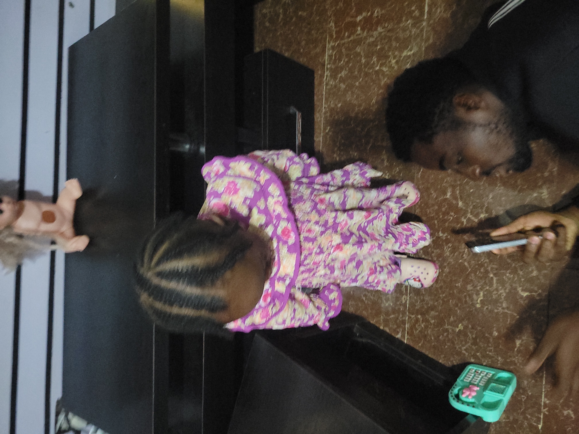 In an indoor setting, a young child wearing a floral dress is seen from a high angle, looking down. The child has braided hair and is wearing light-colored shoes. To the right, an adult male, with dark hair and a beard, is looking at a smartphone he is holding. The floor has a marble pattern. In the foreground left, a teal toy calculator is visible. A dark, rectangular table is in the background, and on top of it, a doll is lying down.