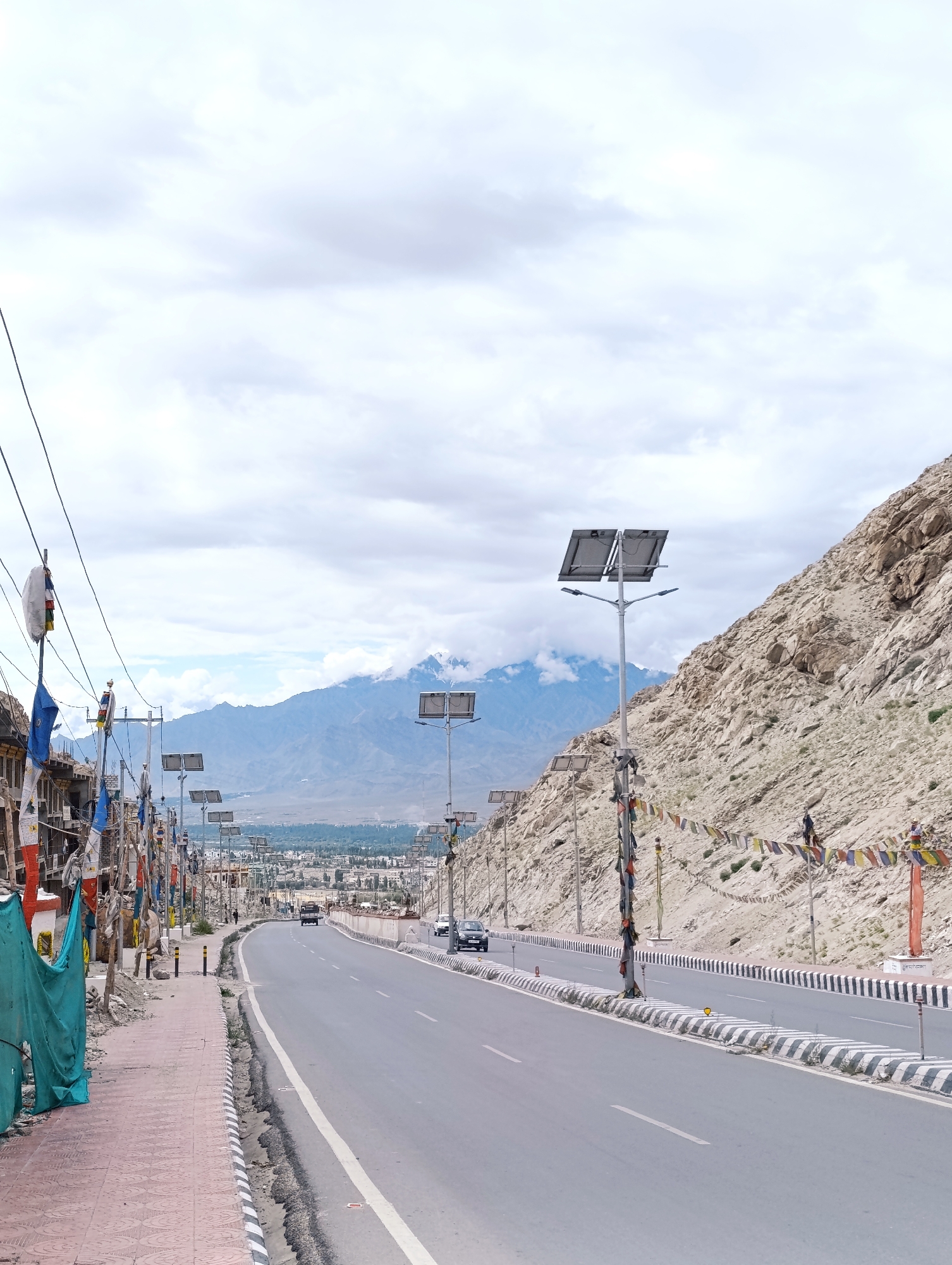 The image shows a paved road leading into a valley in the city of Leh, India. The road is lined with solar-powered streetlights and prayer flags. On the left, a sidewalk with red brick paving is visible, with some buildings and a green tarp. On the right, a steep, rocky mountainside dominates the landscape. In the distance, mountains are visible under a cloudy sky. Several cars are on the road, and a few people are walking on the sidewalk. The scene appears to be set during daytime.