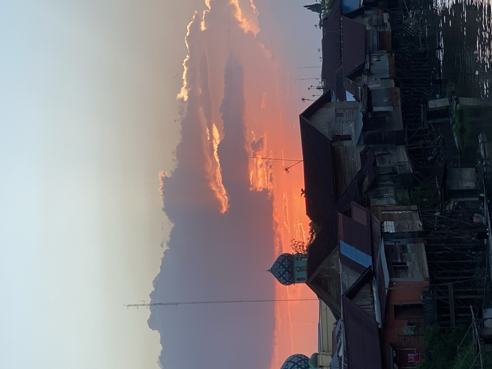 The image depicts a sunset over the city of Tunggul Irang Ulu, Indonesia, with a focus on the stilt houses along a body of water. The sky is a gradient of pale blue at the top transitioning to vibrant orange and pink hues near the horizon, illuminated by the setting sun. Fluffy clouds are scattered across the sky, catching the warm light.
In the foreground, a row of traditional stilt houses with weathered wooden walls and corrugated metal roofs lines the water's edge. Some of these houses have visible occupants or evidence of daily life, such as laundry hanging out to dry. The stilt structures are a prominent feature, suggesting the houses are built over the water or marshland. A river or canal flows in front of the houses, reflecting the sky and the structures.
Further back, the domes and minarets of what appear to be mosques rise above the rooftops, characteristic of the local architecture. A tall radio tower stands prominently against the sky on the left side of the image. The overall scene conveys a sense of community life and the unique architectural style adapted to the local environment.