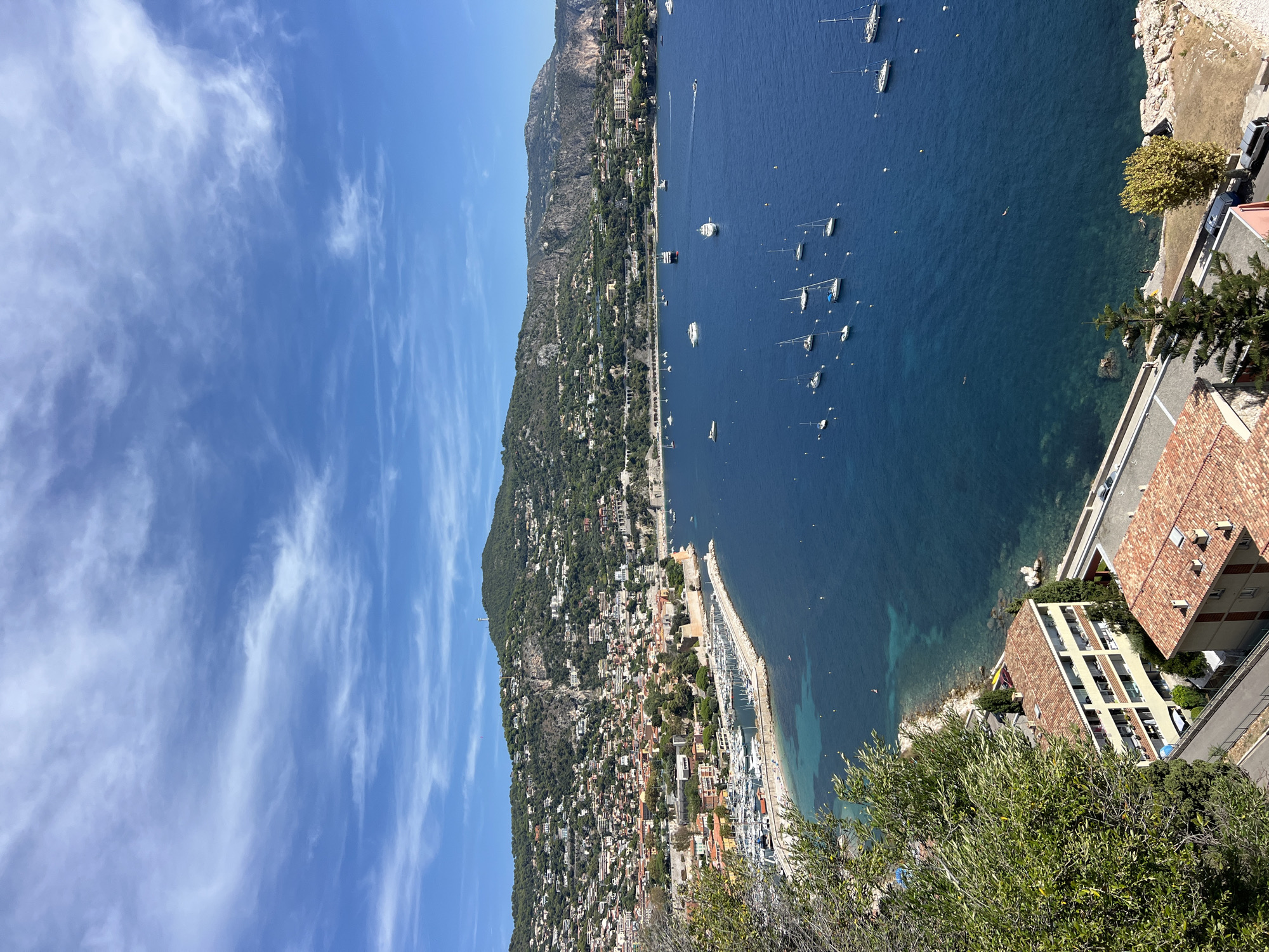 This is a high-angle, daylight photograph of a coastal town nestled against a lush, green hillside. The scene is dominated by the deep blue of the Mediterranean Sea, dotted with numerous sailboats and a few larger vessels. In the foreground, a curved marina wall is filled with docked sailboats. Beyond the marina, the town sprawls up the hillside, characterized by terracotta-tiled roofs and a dense collection of buildings. The hillside itself is covered in trees and vegetation, with a winding road visible in the lower portion of the image. The sky is a vibrant blue, streaked with thin, white clouds. The overall impression is one of a picturesque, sunny day in a Mediterranean coastal resort town, likely somewhere along the French Riviera, such as Nice, France, given the architecture and the style of the coastline and hillside development.