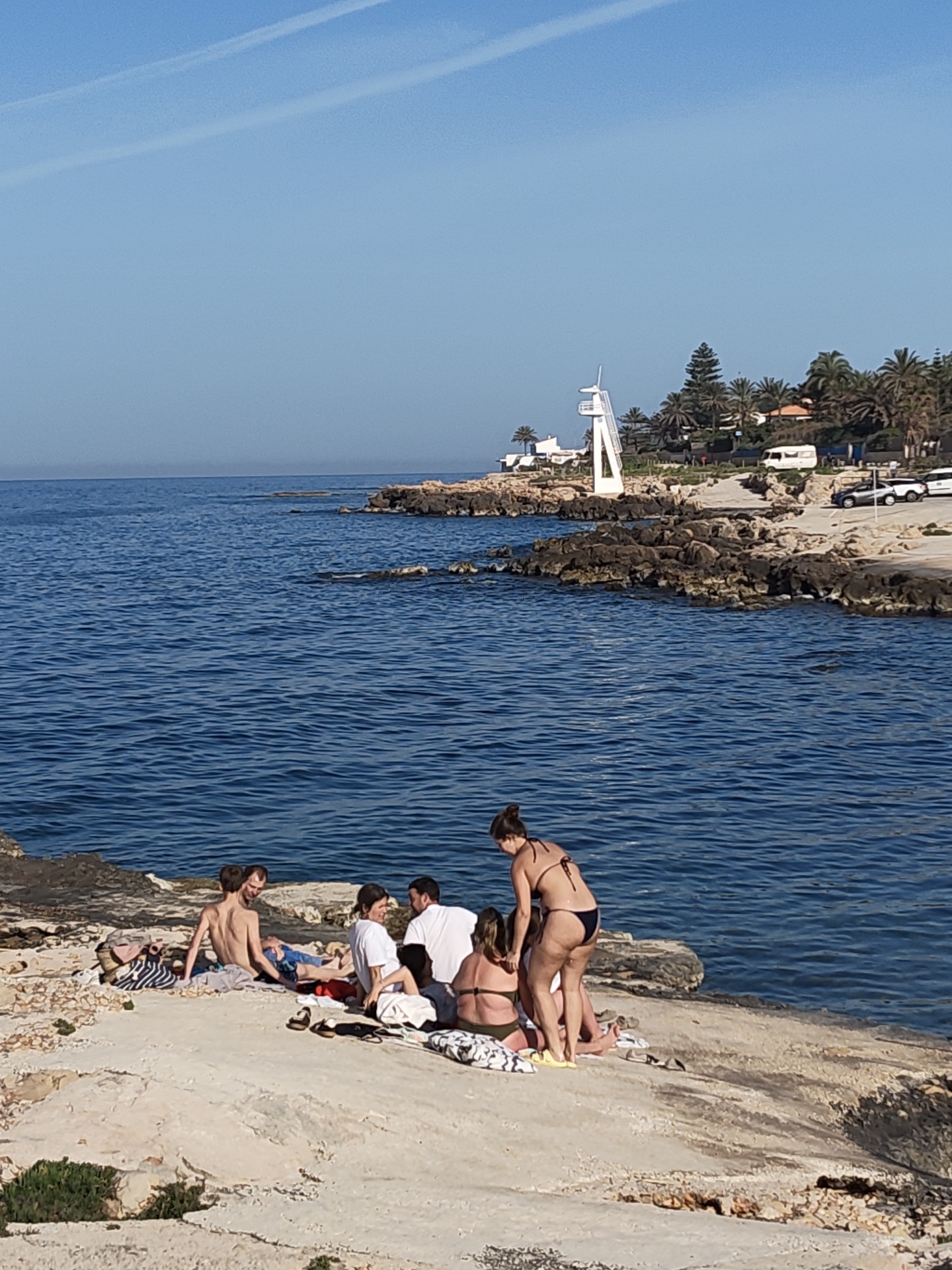 A daytime shot in Rotes, Spain, shows a group of seven people on a rocky, seaside location. To the left of the frame, two men are sitting, one shirtless and the other in blue swim trunks. In the center, four individuals are gathered, with three seated and one woman standing in a black bikini. A man is seated in a white shirt. To the right, a woman in a bikini is assisting another woman who is seated. The rocky terrain extends into the ocean. In the background, a white lifeguard tower is visible near parked cars, palm trees, and other vegetation. The sky is clear.