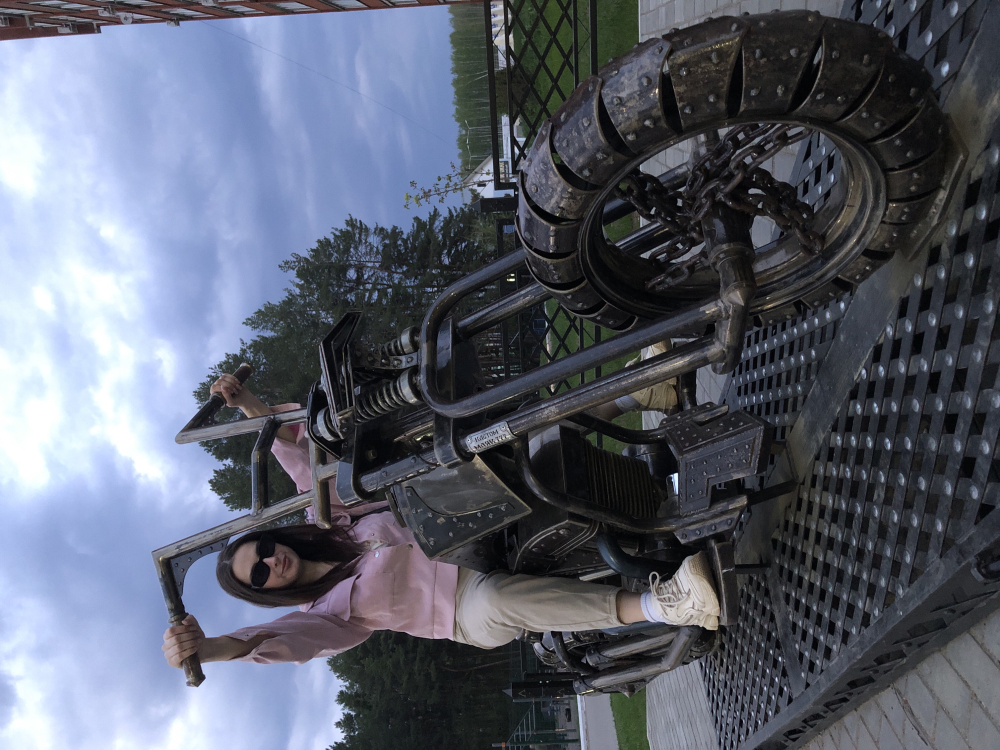 A woman is posing on a motorcycle sculpture in Chaikovsky, Russia. The motorcycle is metallic, with a large, segmented tire held in place by a chain. She is wearing a light pink shirt, light khaki pants, white sneakers, and sunglasses. The sky is overcast. Trees and a building can be seen in the background. The sculpture rests on a raised platform with a patterned metal surface.