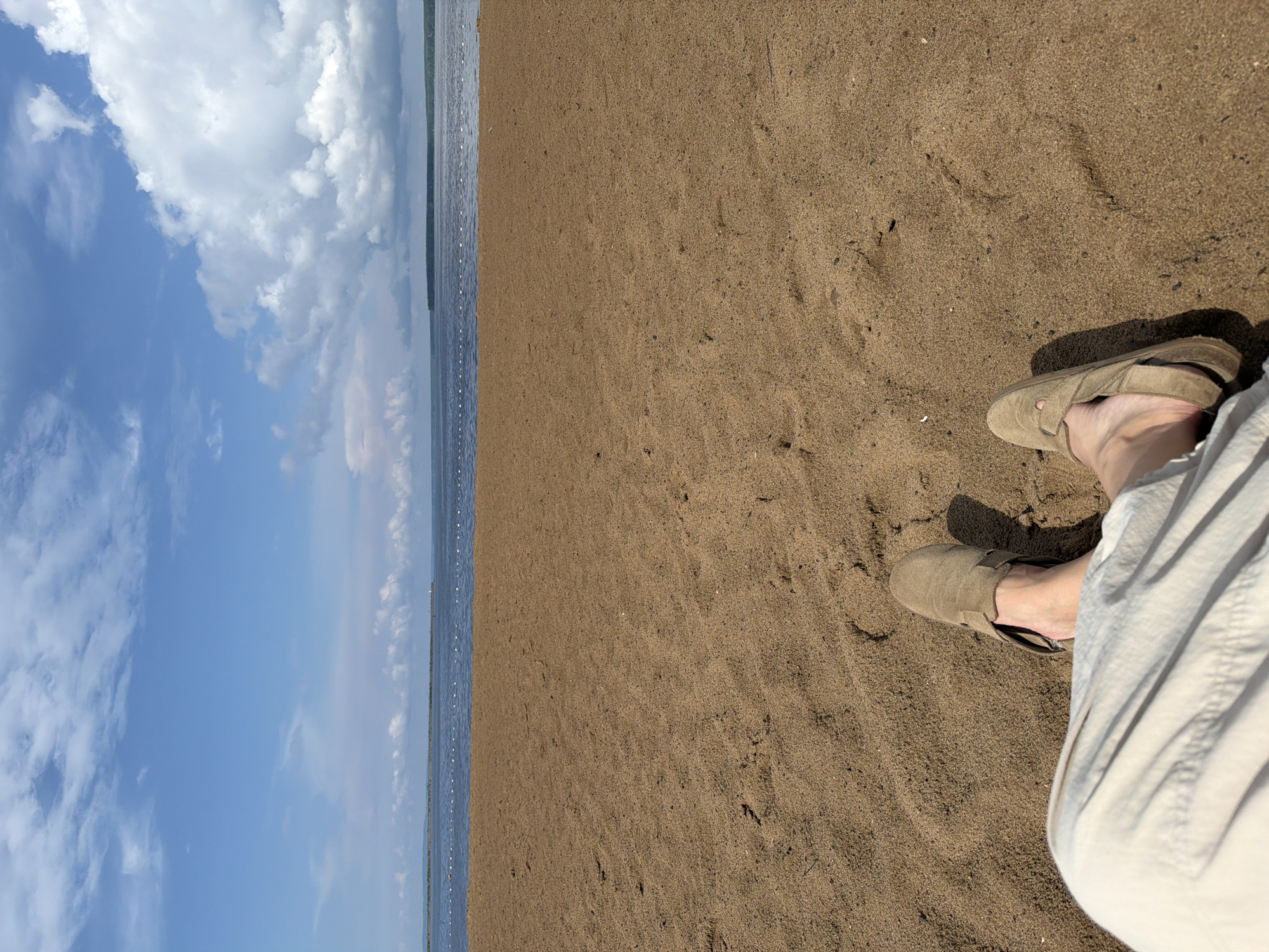 A first-person perspective shot captures a pair of feet, clad in beige sandals and light-colored pants, resting on a sandy beach. The sandy expanse dominates the foreground, with gentle undulations and scattered debris. Beyond the beach, a calm body of water stretches to the horizon, marked by a line of buoys. The sky above is a vibrant blue, dotted with fluffy white clouds. The scene is likely located on the banks of the Kama River in Chaikovsky, Russia, given the characteristic landscape.