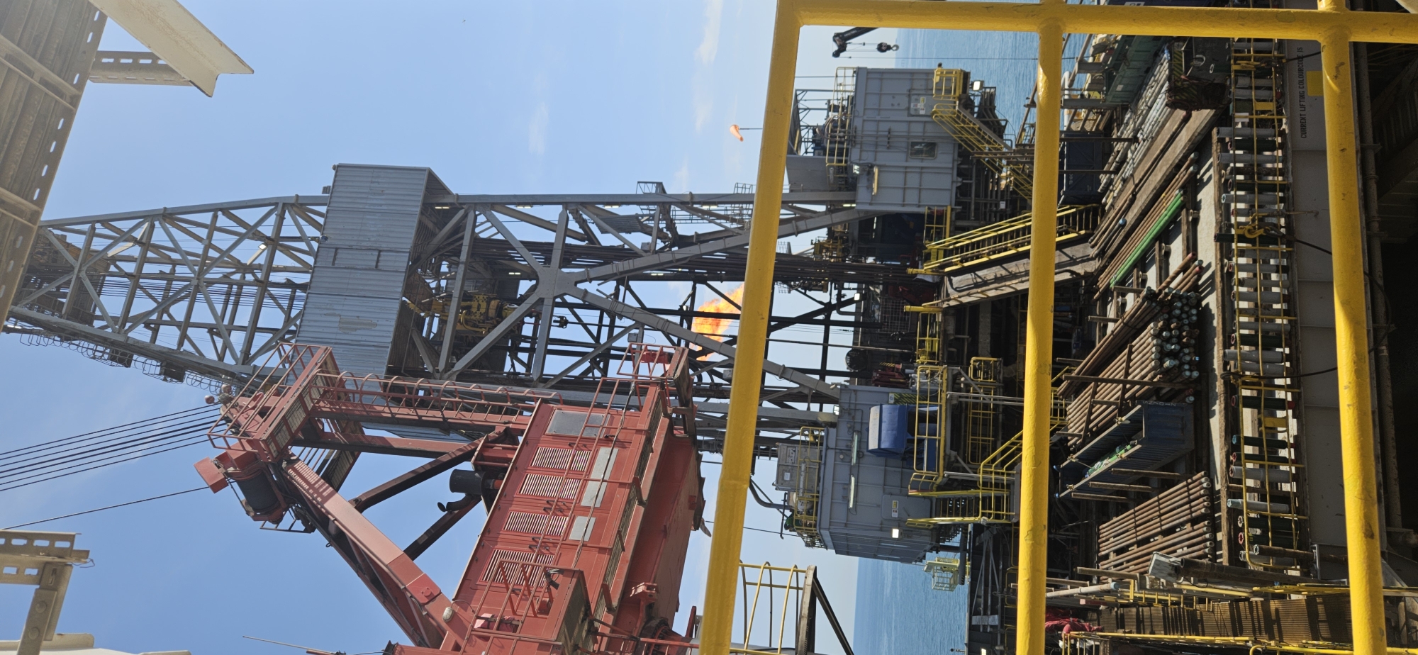 An offshore oil rig is depicted under a clear blue sky. The rig is a complex structure of metal beams, scaffolding, and machinery, dominated by a tall derrick. A red crane arm extends from the left side of the frame. The platform below the derrick is cluttered with pipes of various sizes, some bundled together and others standing upright in racks. Yellow safety railings are prominent in the foreground. In the background, the blue ocean stretches to the horizon. A flare stack on the rig is emitting flames. The scene appears to be an industrial environment focused on oil extraction or processing at sea.