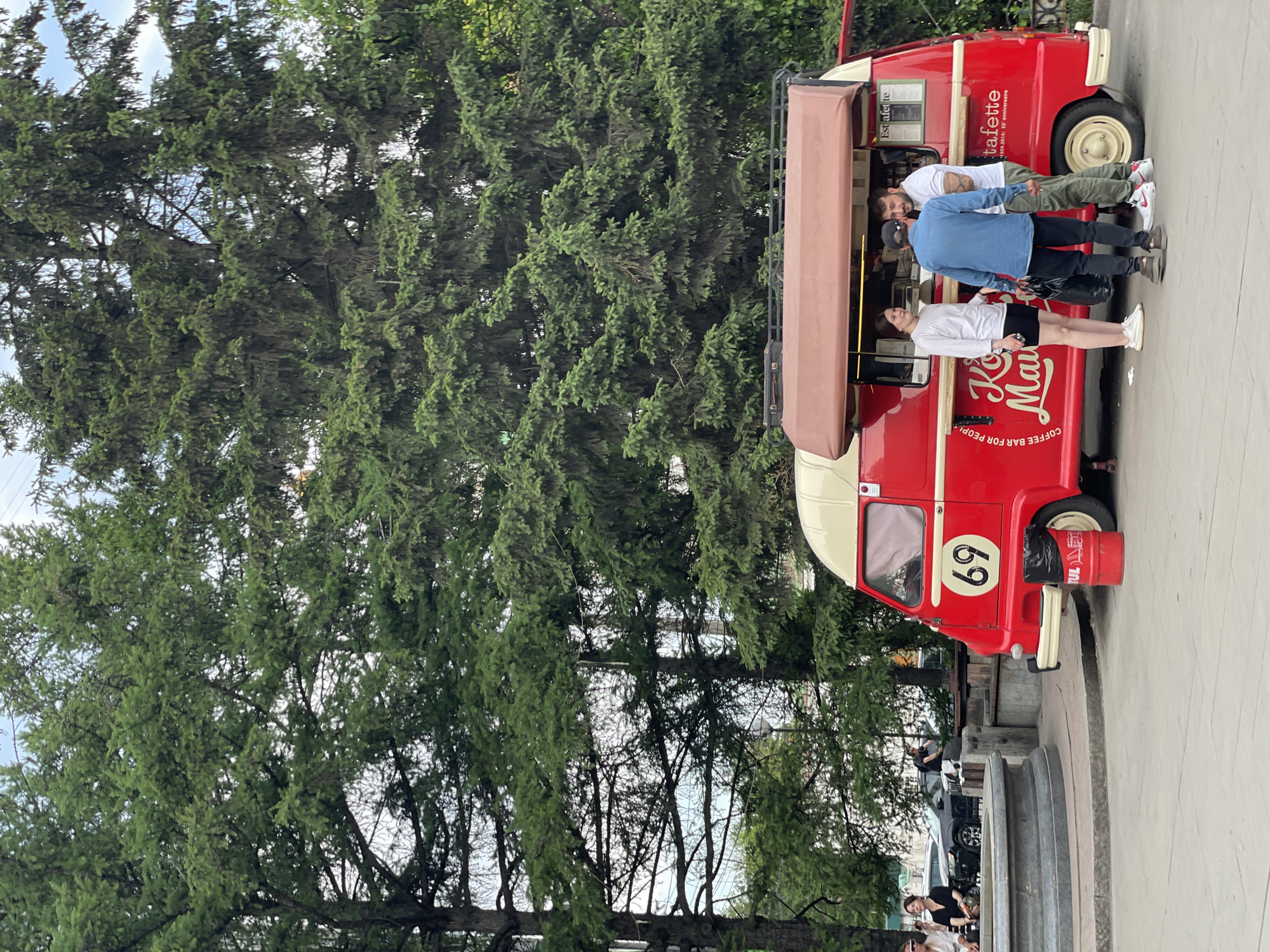 The image depicts a red food truck with a beige awning parked in a paved outdoor area, likely a public square or park. Large evergreen trees form a dense backdrop. Several people are gathered around the food truck. A young woman in a white long-sleeved shirt and black shorts stands to the left of the service window. Two men are conversing in front of the window. One man wears a blue denim shirt and dark pants, holding a black plastic bag. The other man wears a white t-shirt and camouflage-patterned pants. A red trash bin with a black lid is placed next to the food truck. In the background, a few other individuals are seated near a concrete fountain structure. A black SUV is parked behind them. The food truck has the number "69" in a yellow circle on its side, and text that reads "Kof." and "tafette". The scene appears to be an outdoor commercial area with recreational elements. This scene is located in the city of Krasnoyarsk, Russia, specifically on Teatralnaya Square, near the Monument to the First Settlers of Krasnoyarsk.