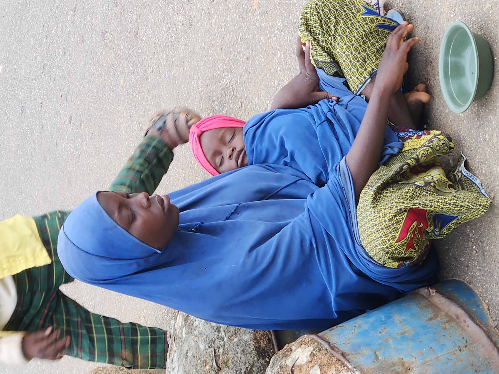 A medium shot shows a young Black woman wearing a blue hijab and a patterned skirt sitting on the ground. Her eyes are closed, and her head is tilted down. Next to her, a baby wearing a pink turban sleeps, cradled in her arms. The woman's right hand rests on the baby, while her left hand is extended towards a small green bowl placed on the ground in front of her. To the left of the woman, a portion of a large, rusty blue barrel is visible. In the background, the legs of another person are visible, wearing green and plaid pants and sandals. The ground is an asphalt surface.
