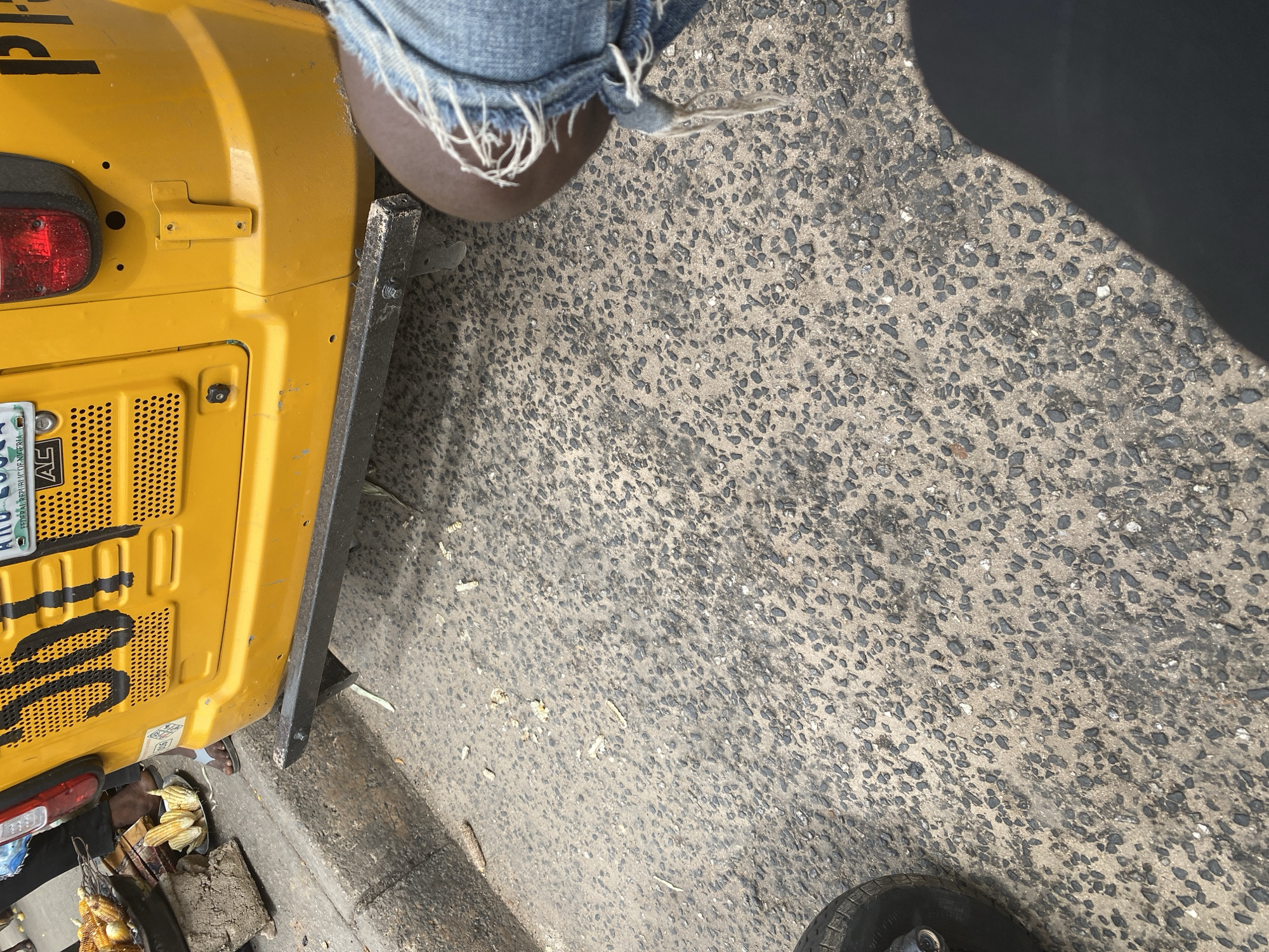 In Jalingo, Nigeria, on a paved street, a yellow auto-rickshaw with the number "500" painted on its side is parked. The auto-rickshaw has a license plate that reads "NG 2958 L." In front of the auto-rickshaw, corn is being roasted on a grill, and several ears of corn are displayed on a plate. A person's legs, wearing distressed denim shorts, are visible in the foreground. The ground is covered in small, dark pebbles and scattered debris, suggesting a busy urban environment. A shadow falls across the lower right portion of the image.
