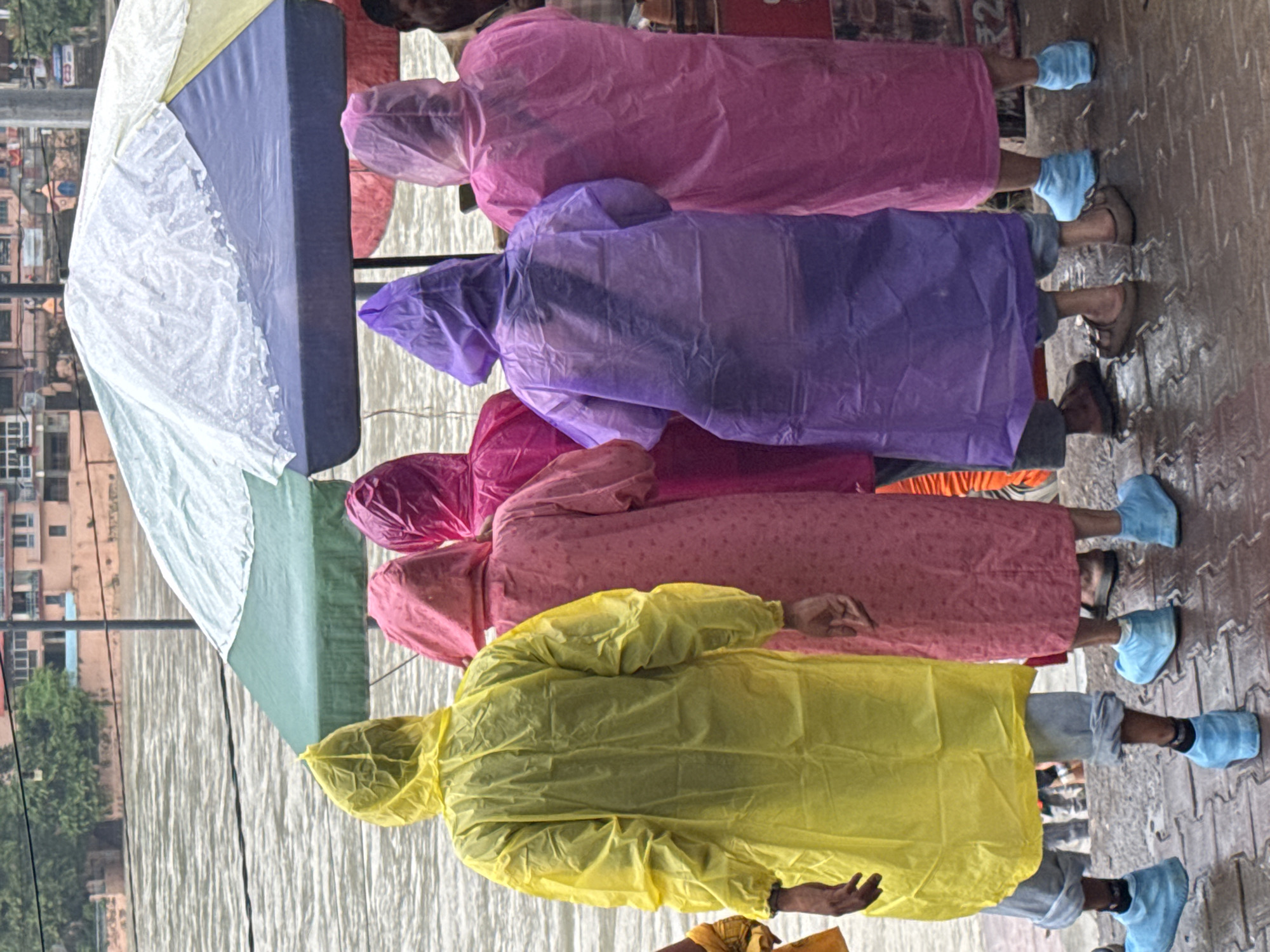 In Rishikesh, India, a group of individuals stand facing away from the camera near the Ganges River. They are wearing brightly colored, disposable raincoats in yellow, pink, and purple. Some individuals are also wearing blue disposable shoe covers. They are standing on a wet, brick-patterned pathway. Overhead, a large, multi-colored umbrella is partially visible, providing shelter. The river is visible in the background, with buildings and greenery on the opposite bank. The scene suggests a rainy day or recent rainfall, with the atmosphere appearing damp and overcast.