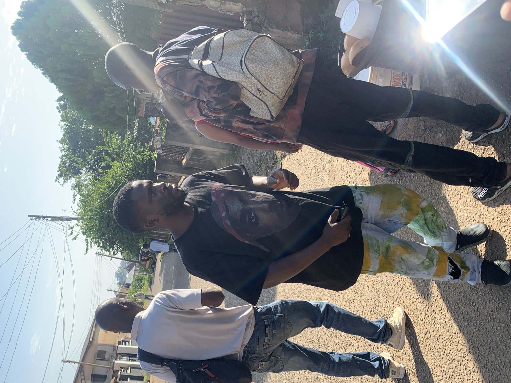 In Jos, Nigeria, a group of young men are gathered on a sunlit, paved street lined with buildings and trees. The image focuses on three individuals in the foreground. The man on the left, wearing a white t-shirt and blue jeans, carries a black backpack. To his right, a man in a black t-shirt featuring a portrait of Tupac Shakur and colorful painted jeans stands with a phone in his hand. Another man, facing away from the camera, wears a tie-dye hooded sweatshirt and a patterned backpack. Behind them, the street continues with a few more individuals and a three-wheeled auto-rickshaw visible in the distance. The scene suggests a casual, everyday encounter on a street in Jos.
