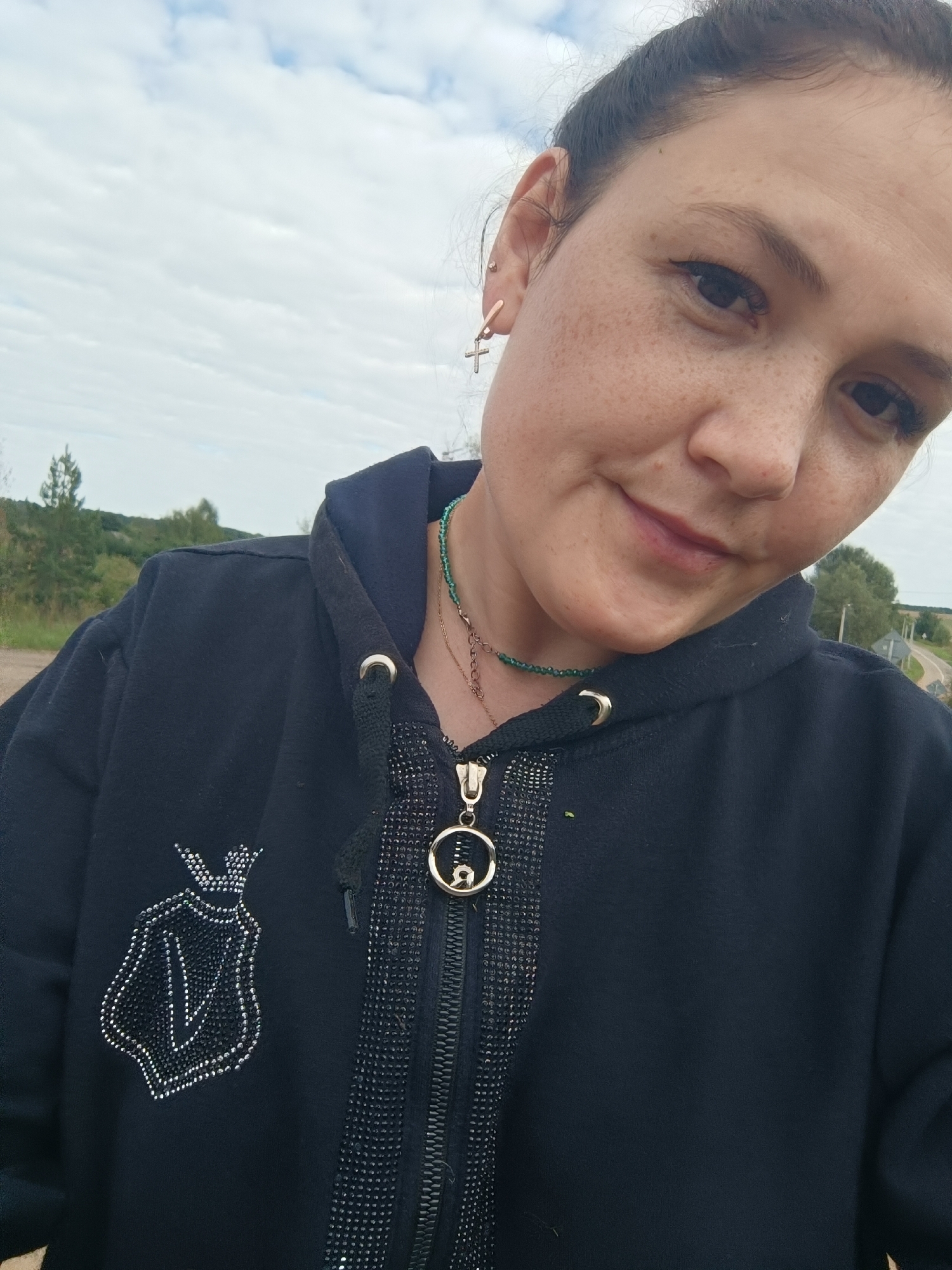 A mid-shot captures a woman from the chest up, facing the camera outdoors. She has dark hair pulled back and freckles across her face. She wears a dark hooded jacket adorned with a beaded crest on the left side of her chest and a zippered placket embellished with black rhinestones. A delicate necklace with small green beads and a chain with a circular pendant is visible around her neck. The background suggests a rural or suburban setting with trees and a cloudy sky. There is no indication of the location being the city of Karsha, Russia.