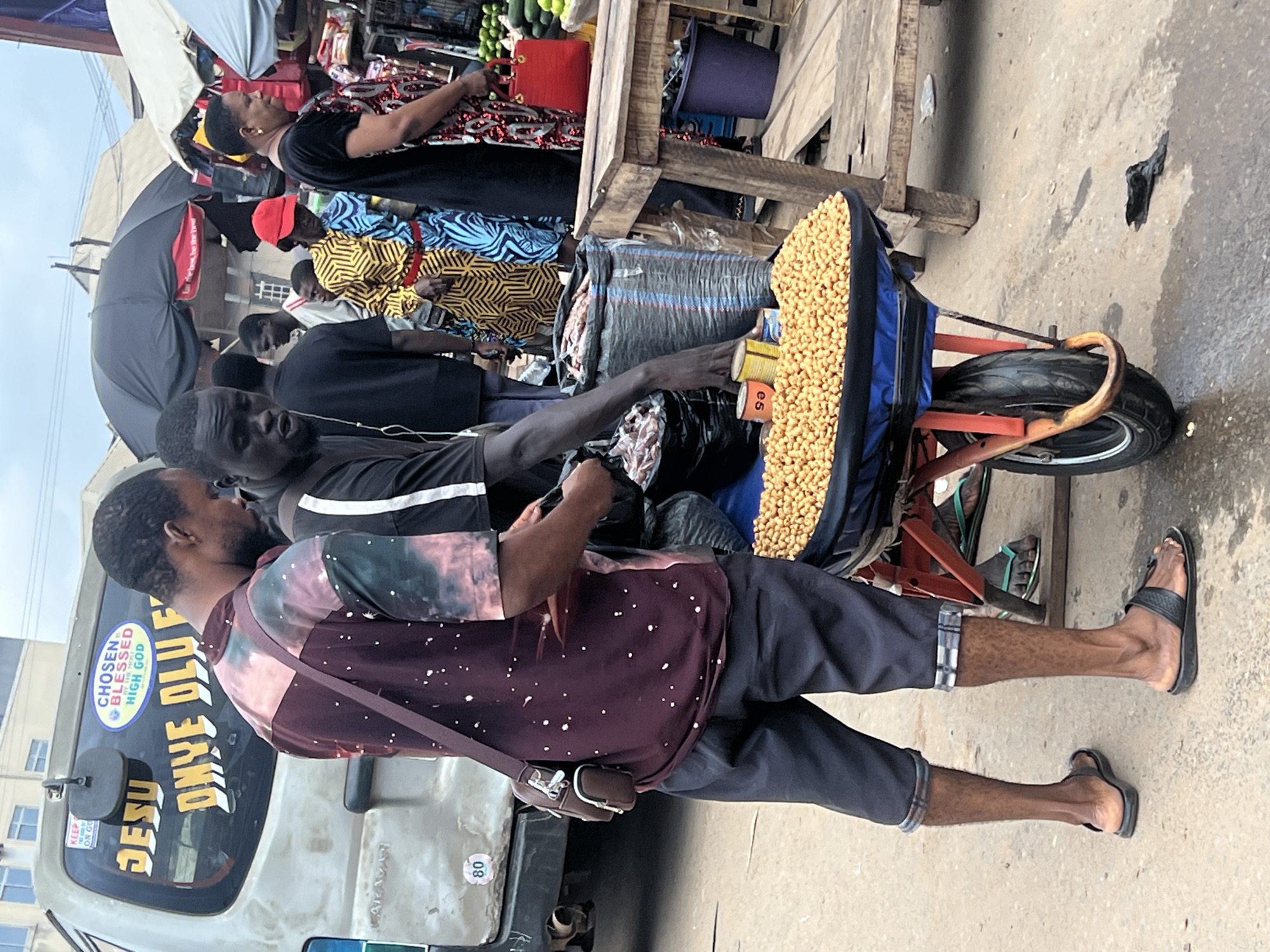 This is a medium shot of a street scene in Nkpor, Nigeria, with a focus on two men interacting near a street vendor's cart. The man on the left, wearing a maroon and pink tie-dye t-shirt and dark shorts, stands with his back partially to the camera, his right hand reaching towards a black plastic bag held by the man on the right. He has a brown satchel slung across his chest. The man on the right, wearing a black t-shirt, is more prominently facing the viewer, his mouth slightly open as he appears to be speaking or attending to the cart. He is holding a black plastic bag filled with goods and is reaching towards a display of small, yellow, bean-like items in a blue-lined cart. Several cans are also visible near the cart, one with visible lettering.
The background reveals a bustling market environment. To the left, a portion of a light-colored van is visible with stickers and text, including "JESU," "CHOSEN," and "BLESSED." Further in the background, a variety of people are seen in colorful attire, some under umbrellas and awnings. A wooden stall with various items, possibly produce like cucumbers and limes, is situated to the right. The ground is paved and appears somewhat wet in places, with some debris scattered about. The overall impression is that of a busy, outdoor marketplace.