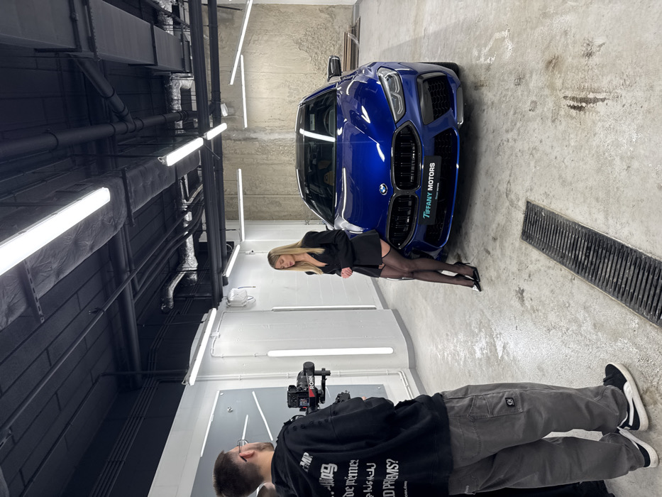 The image captures an indoor video or photo shoot taking place at Tiffany Motors, Lazurowa 27, 01-314 Warsaw, Poland. In the foreground, a male individual, seen from behind, wears a dark hoodie and grey cargo pants while operating a professional camera mounted on a gimbal. To the right, a female model with long blonde hair poses next to a dark blue BMW M8 Competition coupe. She is attired in a black blazer-style top, black thigh-high stockings revealing garters, and black high-heeled shoes. The vehicle's front grille is prominently visible, displaying a license plate frame that reads "TIFFANY MOTORS." The setting features a polished concrete floor with a linear drainage grate, walls composed of both white panels and unfinished concrete, and an industrial-style black ceiling exposing ducts and electrical conduits. Numerous long, bright linear lights illuminate the entire scene.