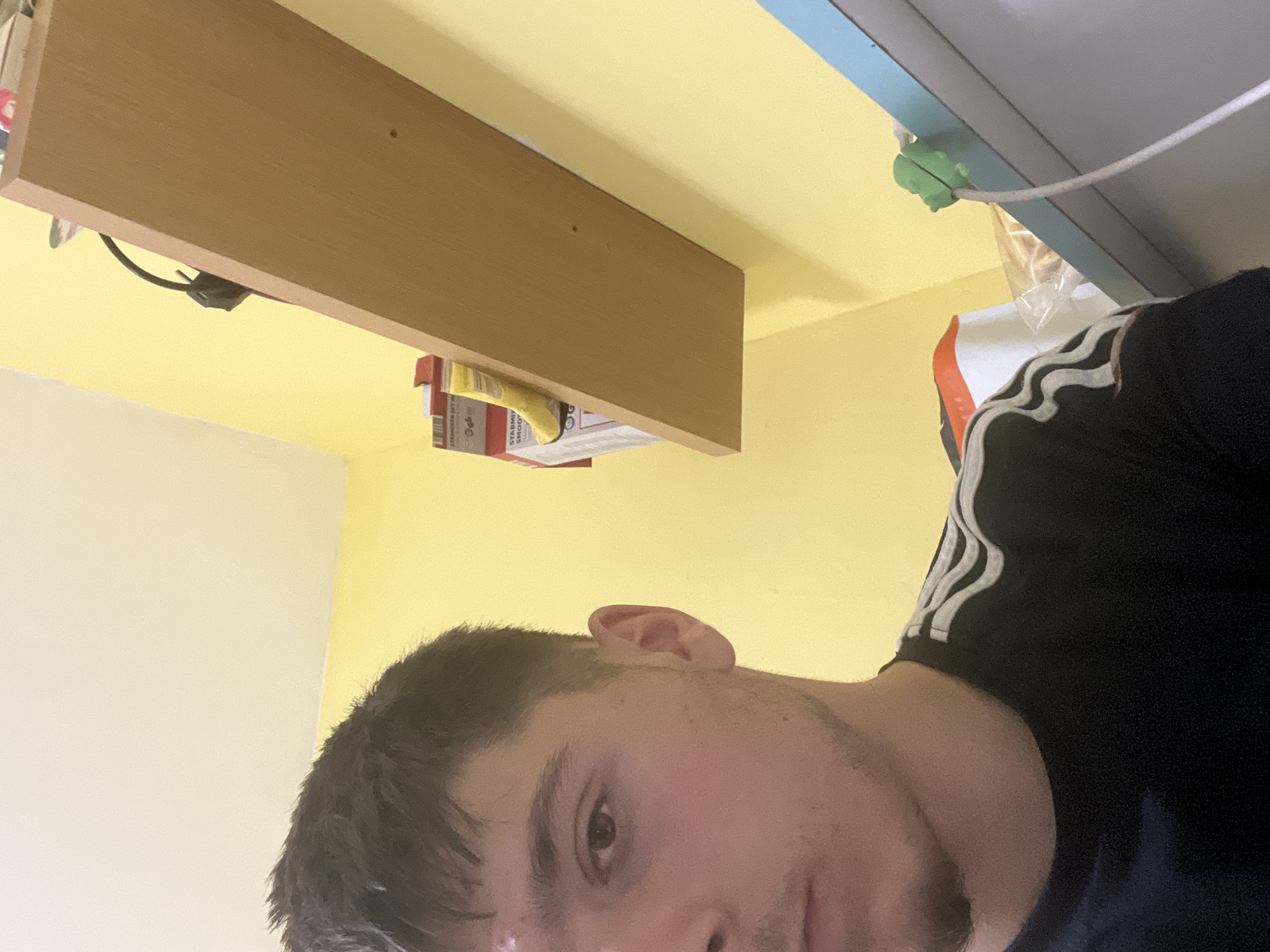 This indoor image is rotated 90 degrees counter-clockwise, showing a young man looking directly at the camera from a low angle. He has short, dark hair and light facial stubble. He is wearing a dark t-shirt with distinctive white stripes on the shoulder, characteristic of the Adidas brand. His expression is neutral.
The setting appears to be a room with vibrant yellow walls. To the left, a section of a white wall or ceiling is visible. Above the man's head, a light brown wooden shelf is mounted horizontally. Underneath the shelf, various items, possibly packages or boxes, are visible, one of which appears to be a "STARBUCKS SET M". A black cable hangs loosely beneath the shelf.
In the upper right portion of the image, a light blue surface or partition is visible, possibly part of a cabinet or furniture. A white cable runs along this surface and is secured by a bright green, frog-shaped cable management clip. Part of a clear plastic bag is also visible near the cable and clip.
The overall scene suggests a casual, personal space, possibly a bedroom or living area. Given the provided context, the location is Burgas, Bulgaria. The time of day is not discernible due to the indoor setting.