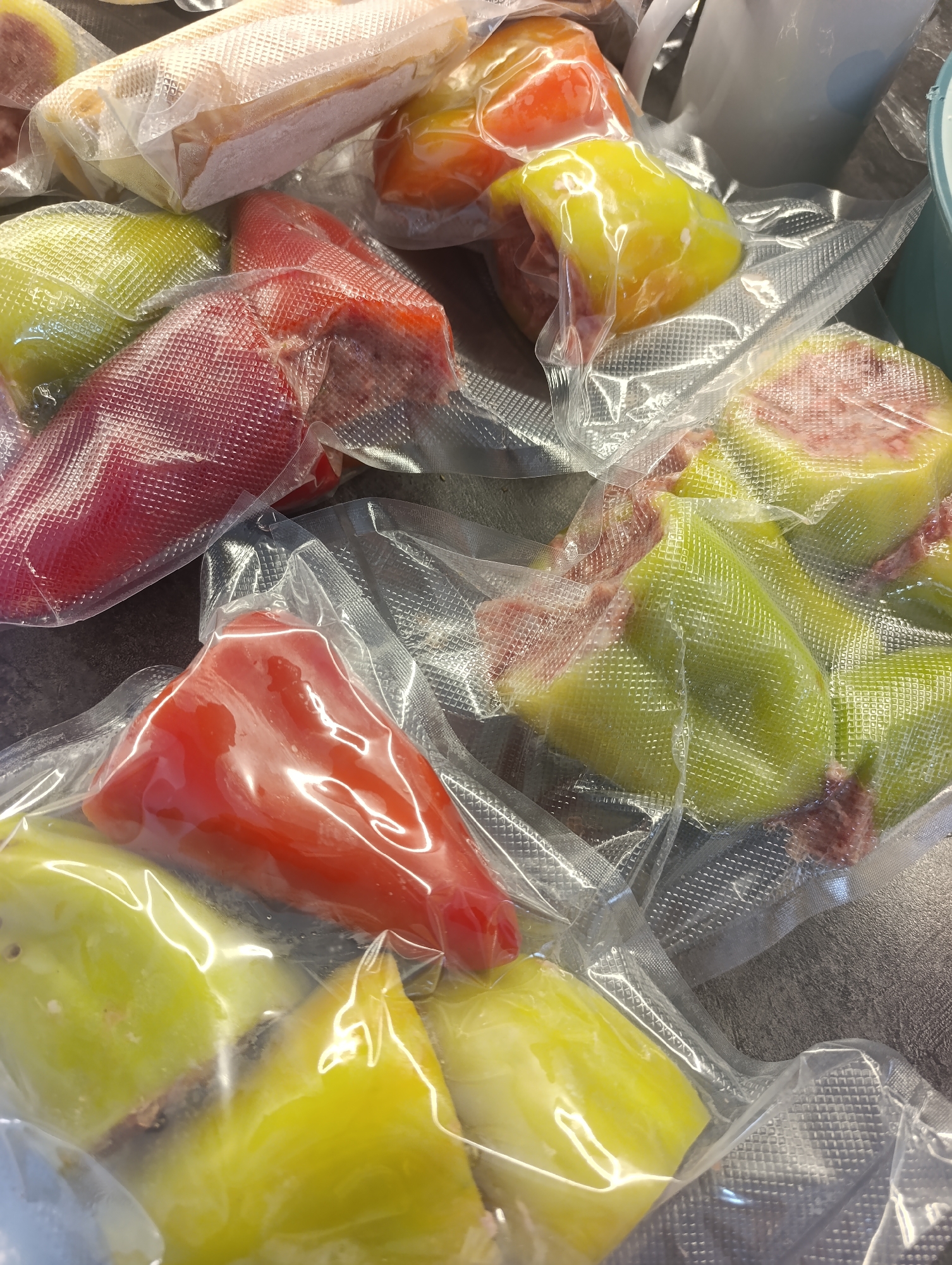 The image is a close-up, top-down view of several vacuum-sealed bell peppers in various colors, including red, yellow, and green. The peppers appear to be whole or halved and are packaged in clear plastic with textured sealing. The background is a dark, textured surface, possibly a countertop. The lighting suggests an indoor environment. There are no people or other identifiable objects present. The location is stated to be Ivanovk, Russia, but this context is not visually discernible from the image itself.