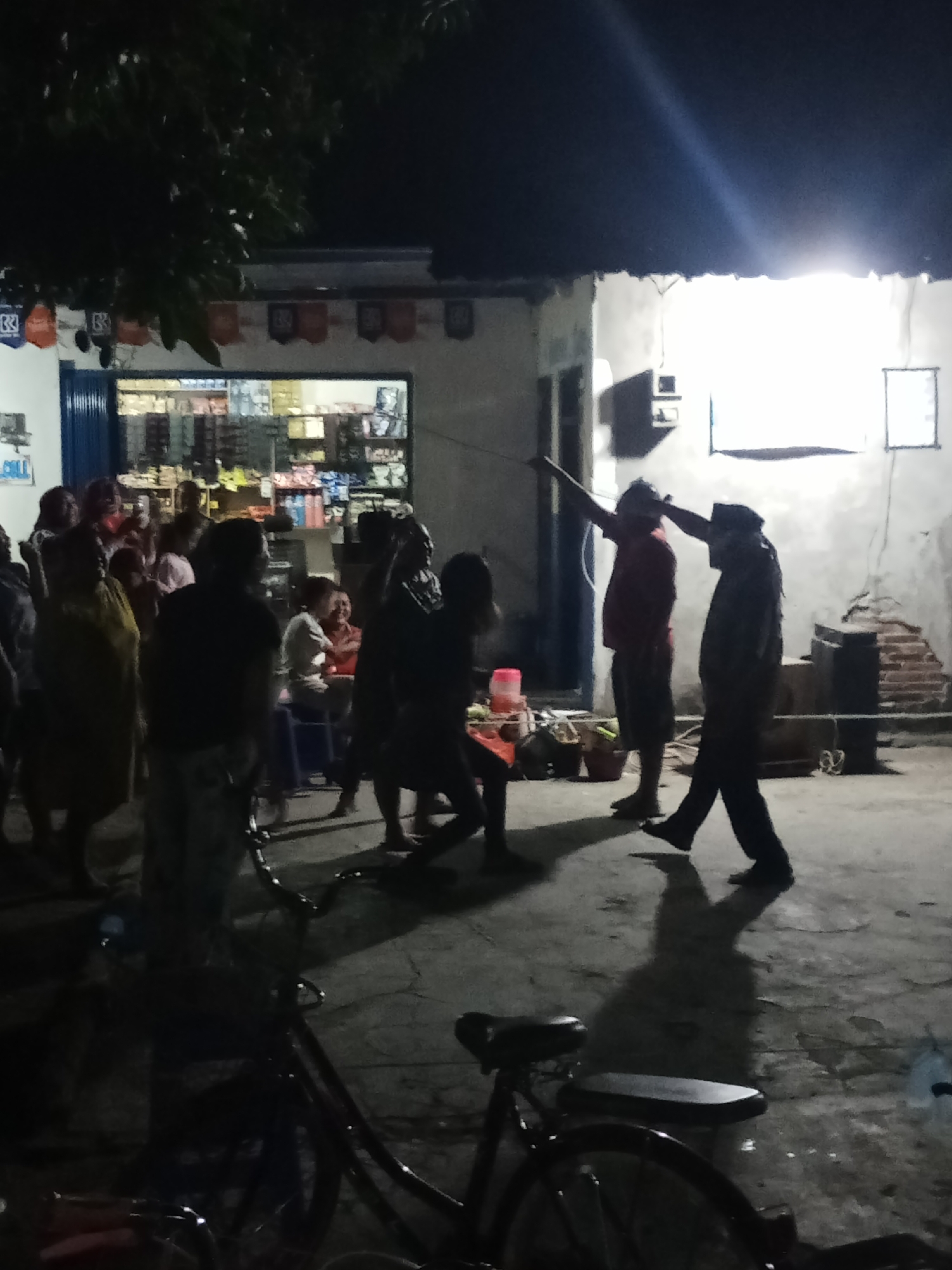 The image depicts an outdoor scene at night in Penggaron, Indonesia, likely a residential area or a small commercial street. In the foreground, a bicycle is parked on a paved surface, casting a shadow. Behind it, a group of people are gathered. A building with a storefront is visible in the background, illuminated by overhead lights. The storefront appears to be a small shop selling various goods, with shelves stocked with items. Several individuals are standing and interacting in the illuminated area in front of the shop. Two men in the mid-ground appear to be engaged in some activity, with one raising his arms. The atmosphere is casual, with people milling around. The lighting suggests it is after dark, with artificial light sources providing illumination.