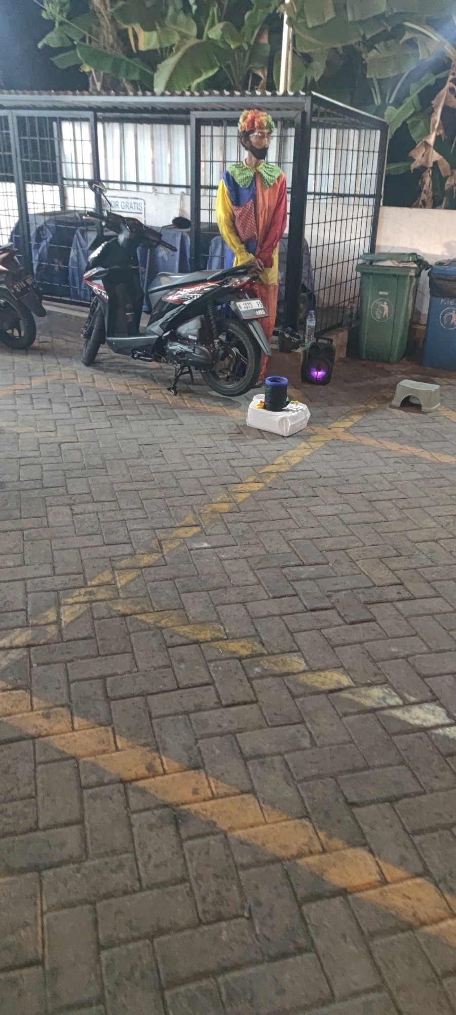 A person in a colorful clown costume stands near a parked motorcycle. The clown has a wig and a ruffled collar. The costume is a patchwork of yellow, red, blue, and green. The person appears to be wearing glasses and a black face mask. The motorcycle is black and appears to be a scooter. It is parked on a paved surface marked with yellow lines. In the background, there are metal fences, trash bins, and lush green foliage, likely banana plants. The scene appears to be an outdoor area in a city, possibly a parking lot or public space. The lighting suggests it is either dusk or an overcast day. The location is Keboansikep, Indonesia.