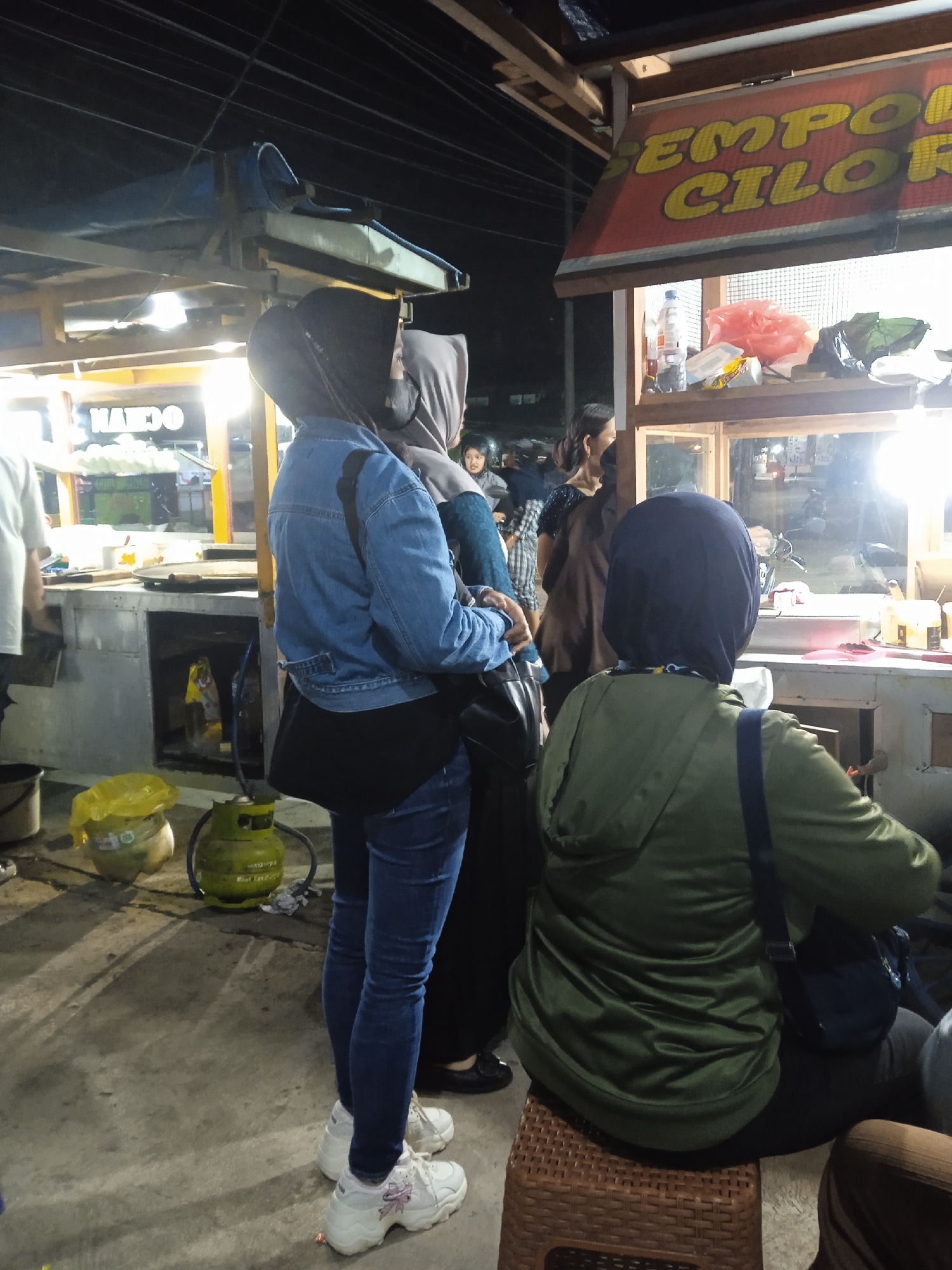 The image captures a vibrant night scene at an outdoor street food market in Kebonsari, Indonesia. Several people, mostly women, are gathered around brightly lit food stalls.

In the foreground, a woman wearing a blue denim jacket, dark jeans, white sneakers, and a black hijab stands with her back partially to the camera, looking towards a stall. She carries a black shoulder bag. Next to her, another woman in dark clothing and a hijab is also visible. To the right, a third woman, wearing a green hooded jacket and a dark blue hijab, is seated on a brown woven plastic stool, facing away from the camera towards one of the stalls.

Other individuals are visible in the background, including a young girl and a person in a white t-shirt on the far left.

Two food stalls are prominent. The stall on the right features a red sign with bold yellow lettering that clearly reads "EMPOR CILOR," indicating it sells a popular Indonesian street food called Cilor. This stall displays various items like plastic bottles and bags. The stall on the left has partially visible text that appears to say "NAKDO" or "KAKO". Both stalls are brightly lit by internal lights, illuminating the surroundings against the dark night sky.

On the ground, next to the stalls, two bright green LPG gas cylinders are visible, typically used for cooking by street vendors. Other items like plastic bags and buckets are also present on the concrete pavement. The scene depicts a casual evening atmosphere where people are likely purchasing or waiting for food.