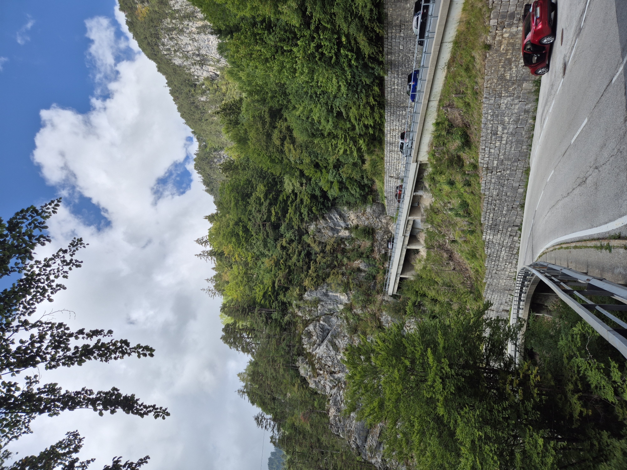 The image depicts a scenic road winding through a mountainous, forested area. The road is paved with white lane markings and is bordered by a metal guardrail on the left. On the right side of the road, a red car with its doors open is parked. Further up the slope, another road is visible, elevated by a concrete structure and lined with a stone wall. Several cars are driving on this upper road. The background is dominated by a steep, rocky mountainside covered in dense green trees. The sky above is blue with scattered white clouds. The foreground shows the lower part of the road curving to the left, with more trees and vegetation visible on the embankment. The scene suggests a remote, natural landscape with well-maintained infrastructure for transportation. The location is identified as being in the city of Ferlach, Austria.