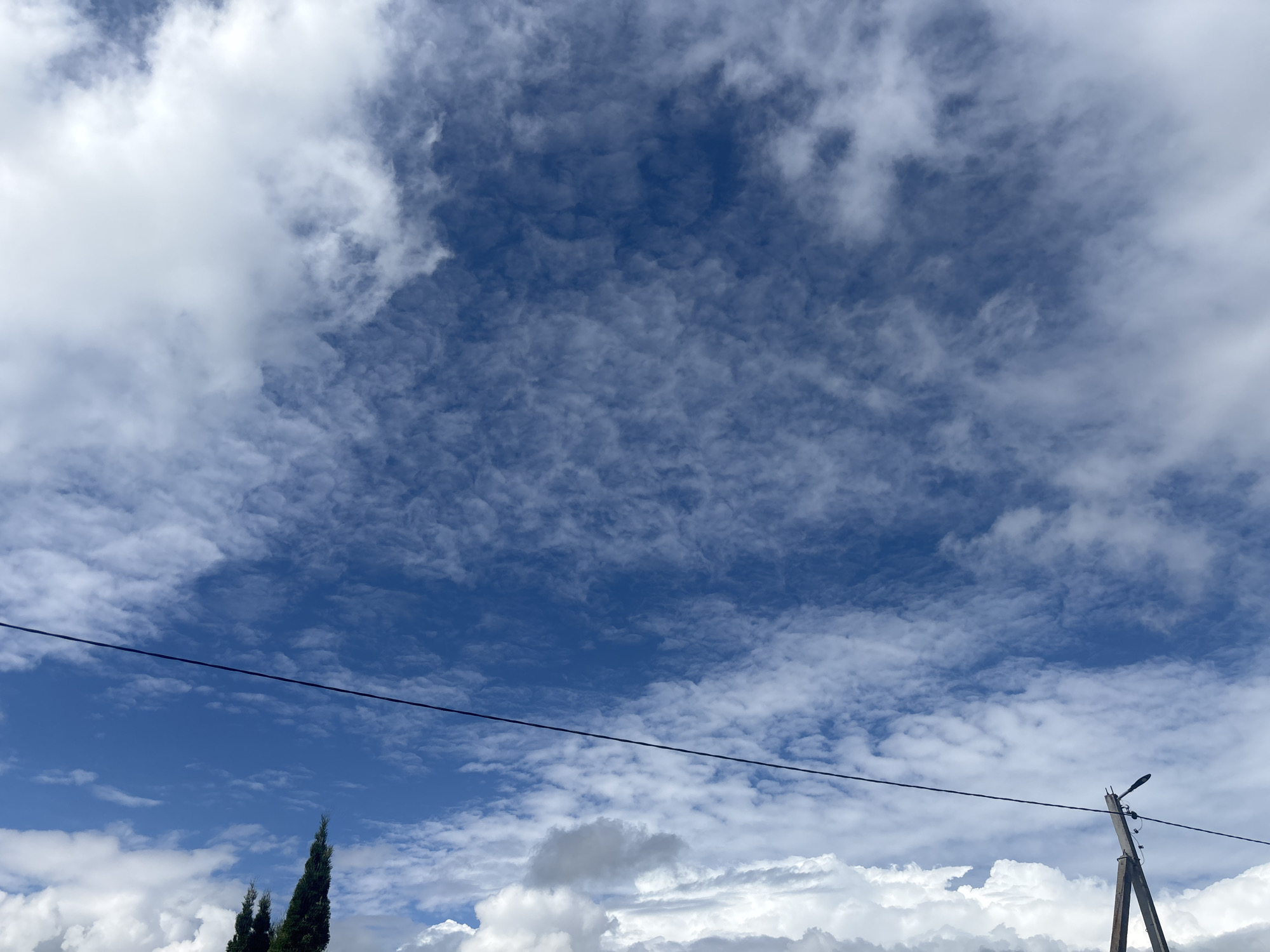 The image shows a cloudy blue sky with some cumulus and cirrus clouds. In the lower left corner, there are a few tall cypress trees. A black power line stretches horizontally across the sky. On the right side of the image, a wooden utility pole with a street light is visible. The scene appears to be outdoors in an open area, likely a rural or suburban setting. There is no indication of the specific location within the city of Kapyĺ, Belarus.