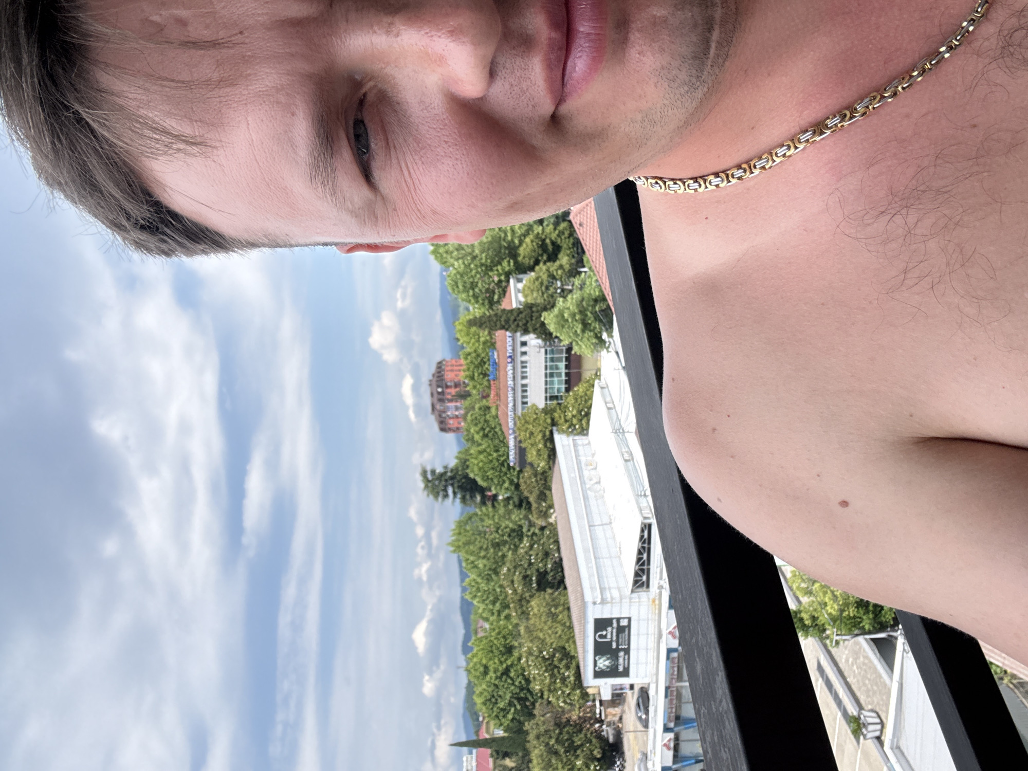 The image shows a close-up of a shirtless man wearing a gold chain, with a blurred background of a cityscape. The city appears to be Sochi, Russia, with buildings, trees, and mountains visible. The man is looking towards the camera with a slight smile. The sky is cloudy. The lighting suggests it is daytime.
