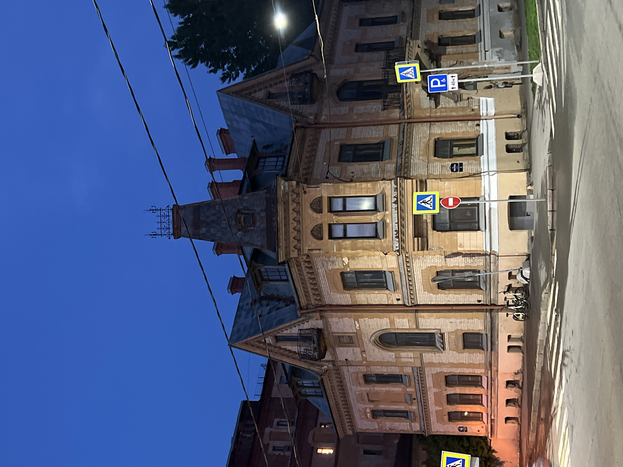 The image shows a corner of an old building in Saint Petersburg, Russia, at dusk or dawn. The building, with a beige brick facade and a dark grey roof, features distinctive architectural elements such as multiple gabled roofs, chimneys, decorative window frames, and balconies. Two bicycles are parked near the building's entrance. Several street signs are visible, including pedestrian crossing signs and a parking sign. The street in front of the building is wet, indicating recent rain, and features a pedestrian crossing. The sky is a deep blue, and the building is illuminated by ambient light.