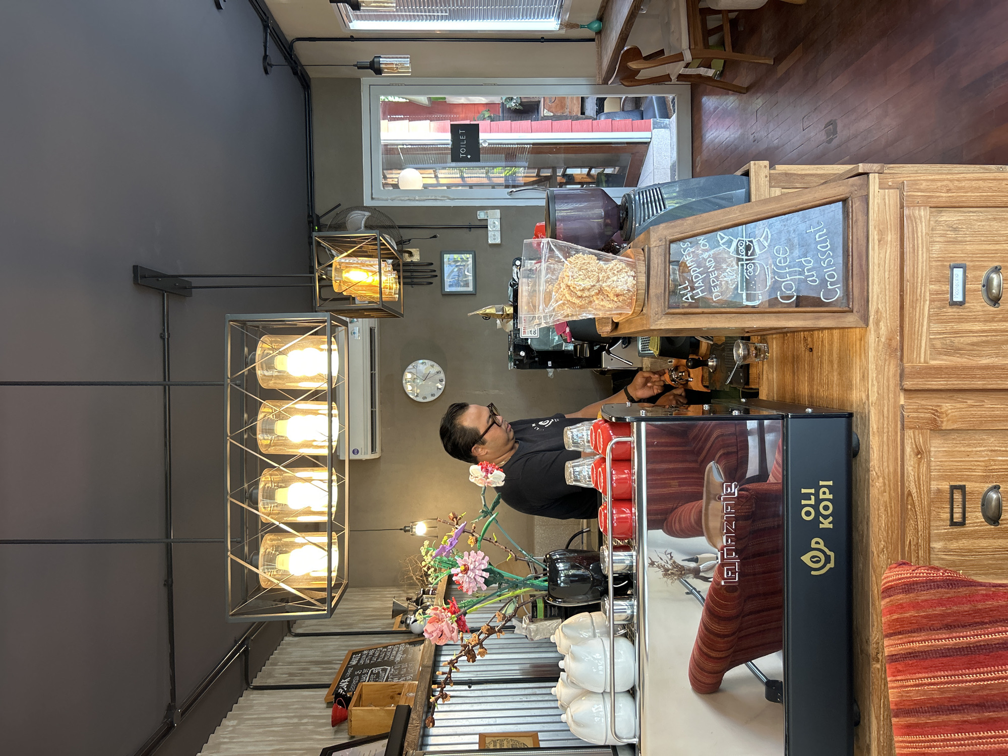 A man in a black t-shirt and glasses stands behind a coffee counter in a cafe. He is wearing a chain with a pendant. The counter is made of wood and features a chrome espresso machine with the brand name "LA PAVONI" and "OLI KOPI" visible. There are several white ceramic jugs and red cups on the counter. A vase with colorful artificial flowers is also present. To the right of the man, a coffee grinder and a clear plastic bag filled with what appear to be crackers are visible. A chalk board sign displays "ALL HAPPINESS DEPENDS ON COFFEE AND CROISSANT." In the background, a door with a "TOILET" sign leads to another area. The cafe has a dark ceiling with industrial-style lighting fixtures. Wooden floors and chairs are visible in the surrounding area. The scene appears to be located in the city of North Kuta, Indonesia.