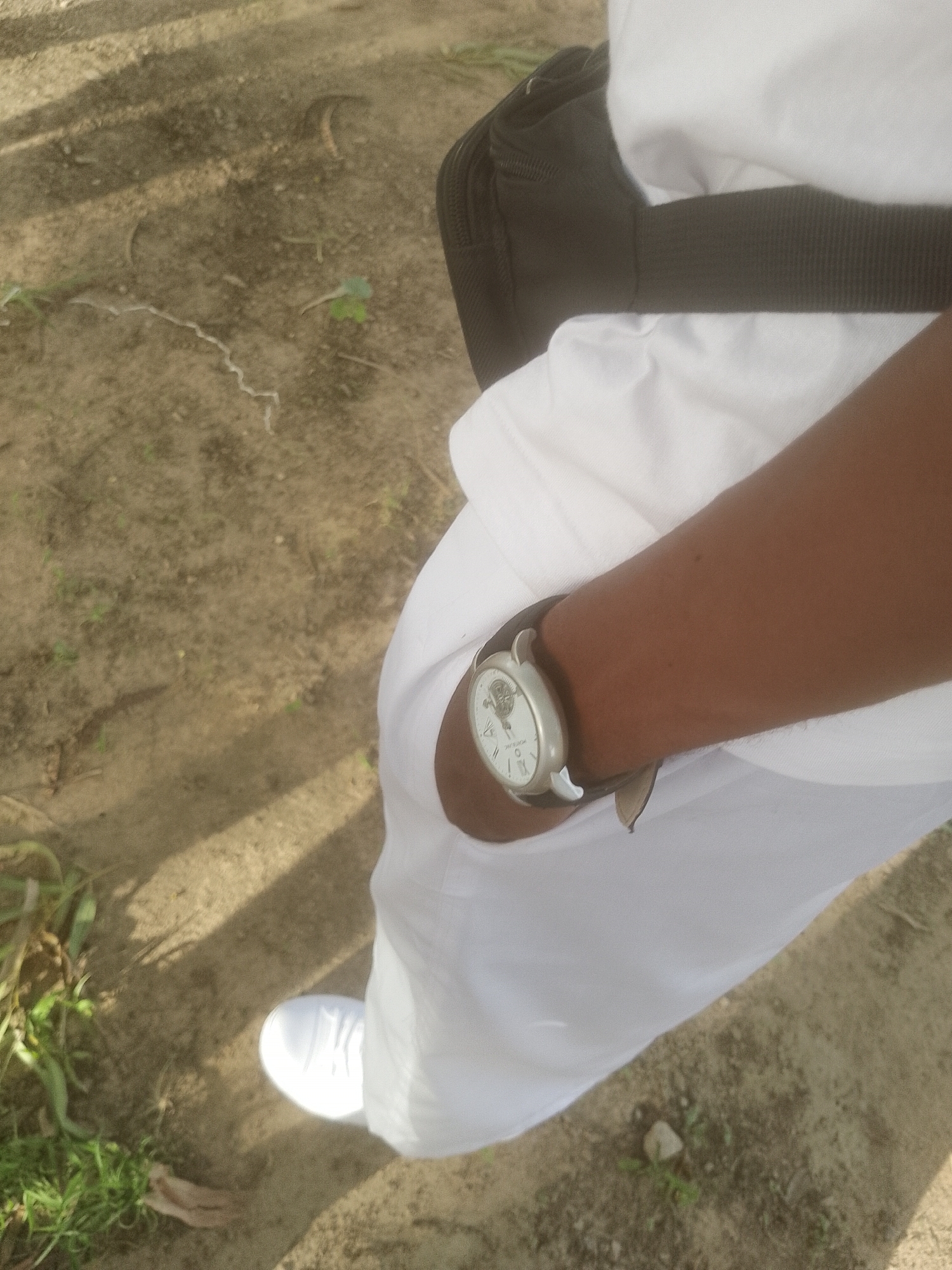 In Majemu, Nigeria, the image captures a close-up view of a person's right arm and lower leg, as seen from a high-angle perspective. The individual is wearing a white garment and a white sneaker. On their wrist, a silver watch with a dark brown leather strap is visible. The watch face is white with a patterned design and Roman numerals for hours. A black strap of a bag is also visible on the person's shoulder. The environment appears to be an outdoor setting with dirt and sparse vegetation, suggesting a casual, possibly rural or undeveloped urban area. The lighting indicates it is daytime, with shadows cast on the ground.