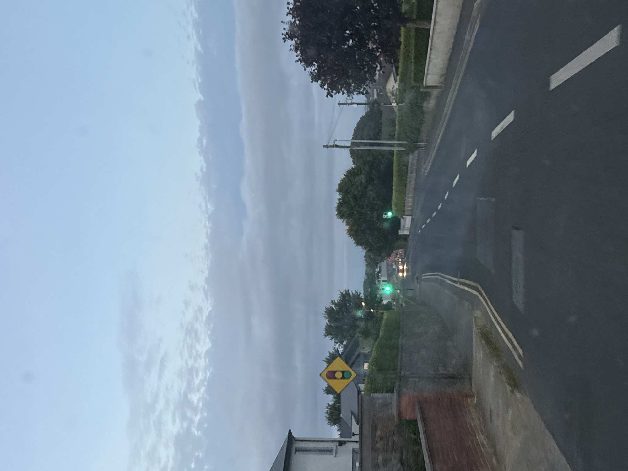 The image depicts a residential street in Tramore, Ireland, likely captured during twilight due to the muted light and overcast sky. The scene is dominated by a road with double yellow lines and dashed white lane markings, suggesting a two-way street. On the left side of the road, a white house with a dark roof is visible, bordered by a low brick wall and a paved walkway. Further down the street, a traffic light is illuminated with a green signal, and a diamond-shaped warning sign displaying a traffic light symbol is also present. Trees and hedges line the sides of the road, with more houses and trees visible in the background. The overall impression is one of a quiet, suburban environment under a cloudy sky.