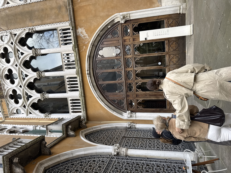 A forensic description of the scene depicts a partial exterior view of the Palazzo Cavalli-Franchetti in Venice, Italy, an edifice characterized by Venetian Gothic architecture. The building facade is an ochre-colored stucco, featuring intricate white stone trim and ornamentation.
The lower level prominently displays a large, rounded archway with a dark wooden double door, elaborately decorated with Gothic tracery, including quatrefoil motifs and pointed arch patterns. To the immediate left of this entrance, another large arched opening is secured by a decorative ironwork grille, flanked by slender stone columns. Above these ground-level features, a balustraded balcony with multiple arched windows (ogee arches, columns, quatrefoil tracery) runs across the upper floor, reflecting the overcast sky and bare tree branches. Sections of the stucco facade show minor discoloration and wear.
On the paved stone ground in front of the building, three individuals are observed from behind, facing the large wooden entrance.
1. **Person 1 (right):** A female with dark hair styled in a bun, wearing a light beige long trench coat and carrying a brown leather shoulder bag. Her left arm rests casually on the shoulder of the adjacent person.
2. **Person 2 (middle):** A female with light-colored, short hair, dressed in a light brown suede-like jacket and white pants. She carries a dark brown or black shoulder bag. Her right shoulder is beneath the arm of Person 1.
3. **Person 3 (left):** A female with dark hair, wearing a black baseball cap and a dark jacket. Only her back and head are partially visible.
To the far left, a white circular table with two white metal chairs suggests an outdoor seating area. To the right of the main wooden door, a tall, slender white vertical signpost with black text and a logo (likely the European Cultural Centre, given context often associated with Palazzo Cavalli-Franchetti) provides information, presumably for an exhibition or event. The overall scene suggests visitors arriving at a cultural institution.