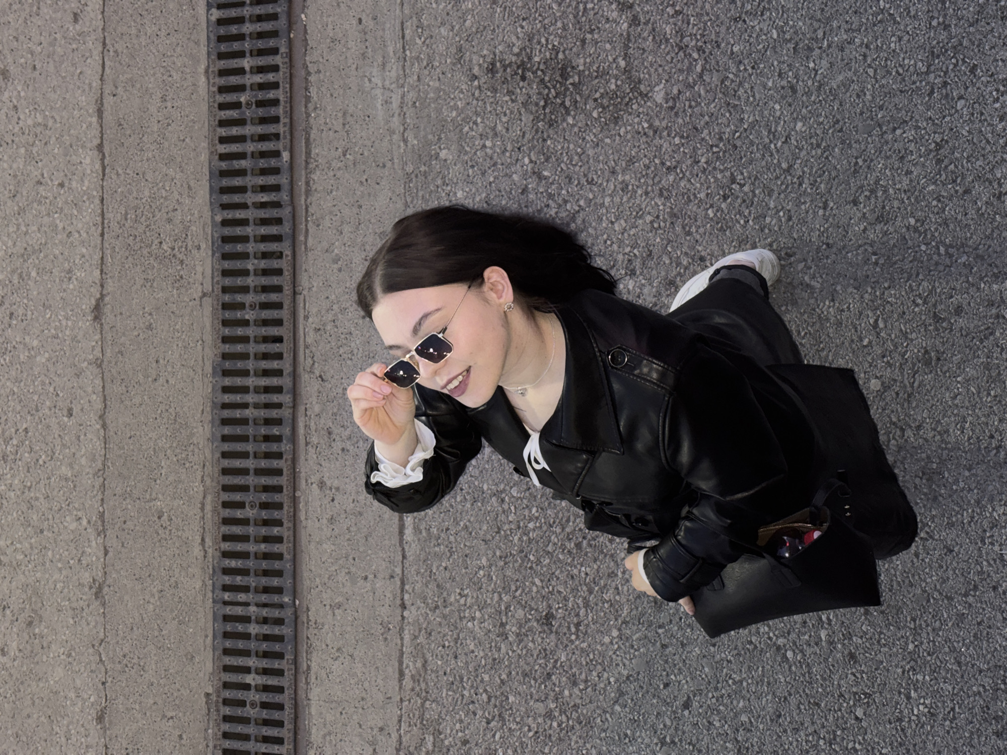 An overhead shot captures a young woman walking outdoors on a textured ground surface, viewed from a high angle looking down. The woman, with dark brown hair and fair skin, is smiling and looking slightly upwards and to her left. She is wearing square-framed, reflective sunglasses, which she is pulling down with her right hand, revealing part of her smile.
Her attire consists of a black leather-look jacket or coat, under which a white top with ruffled cuffs is visible at her wrists and collar. She is also wearing dark trousers and white sneakers on her feet. A delicate silver necklace and small, possibly floral or star-shaped, stud earrings adorn her neck and ears. On her left arm, she carries a black shoulder bag or tote. Her overall expression is one of pleasantness and joy.
The ground she is walking on is divided into two main sections by a long, dark grey metal drainage grate running vertically down the left side of the image. To the left of the grate is a section of lighter grey concrete with a rough texture. To the right of the grate, and covering the majority of the image's right side, is a darker grey asphalt or tarmac surface, also rough and pebbled. The drainage grate itself is long and narrow, featuring numerous small rectangular openings. The lighting appears bright and even, suggesting it is daytime, possibly with overcast skies or diffuse light. No specific location or weather details are overtly apparent beyond being an urban or suburban outdoor setting. There is no visible text in the image.