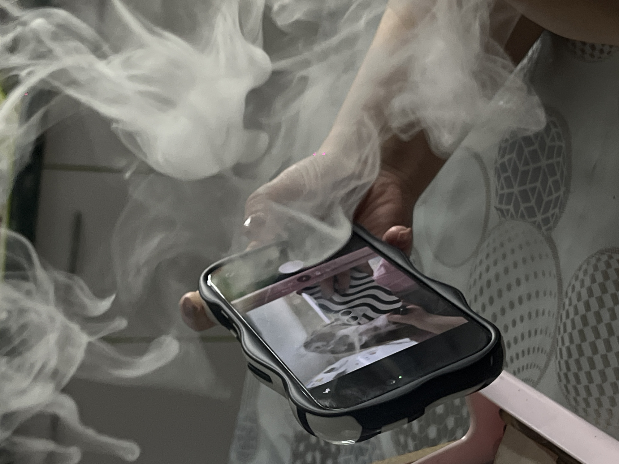 A mobile phone is being held in a hand amidst a cloud of white smoke. The phone is black and is reflecting the image of a cat on its screen. The background features a geometric pattern.
The location of this scene within Port-Katon, Russia, cannot be determined from the visual information available.