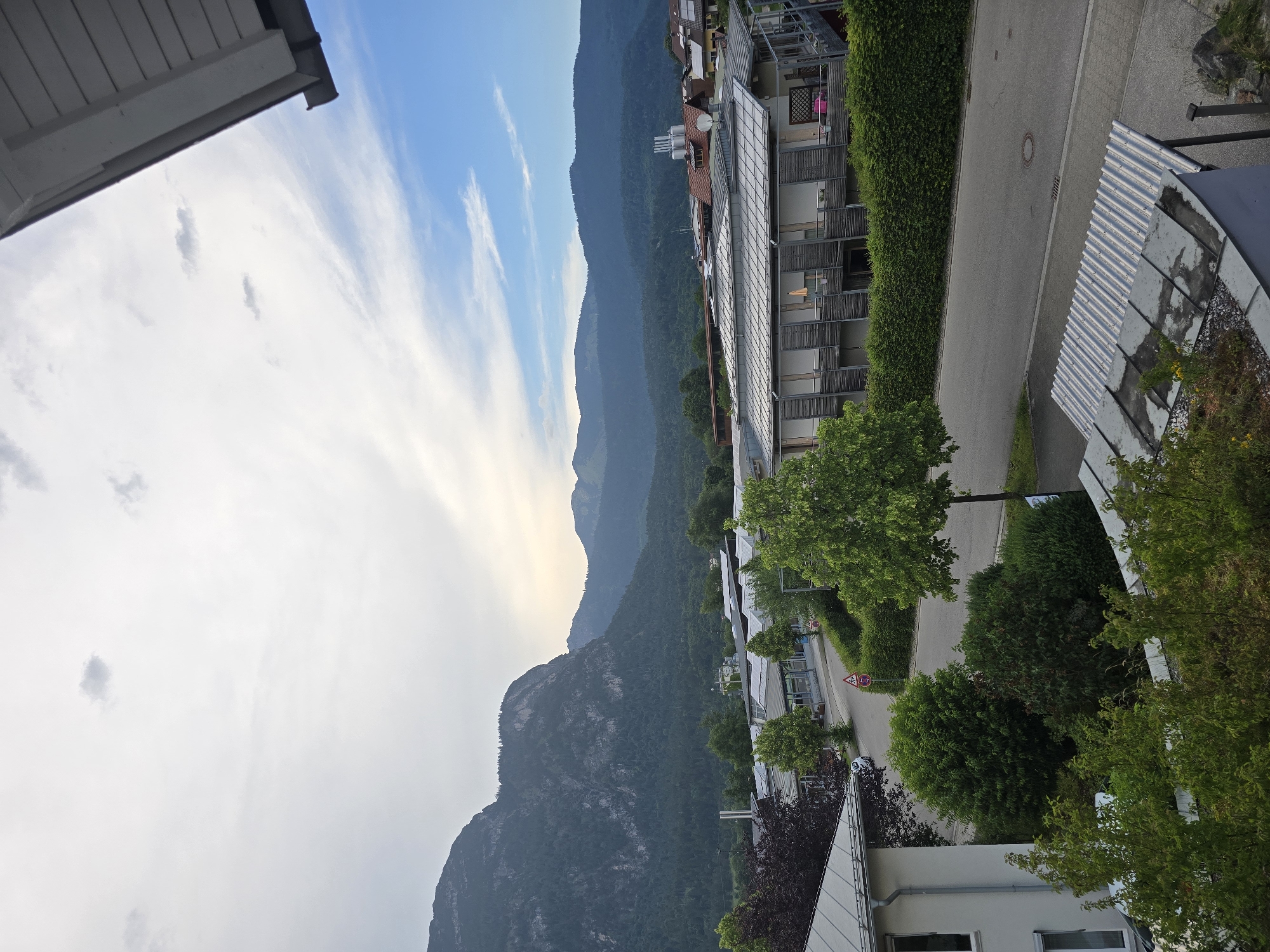 The image depicts a view of buildings and a street with lush green trees and a mountain range in the background. A large, dark-colored mountain dominates the left side of the frame, with a forest covering its slopes and foothills. To the right of this, a range of smaller, more distant mountains forms the horizon.
In the mid-ground, several modern buildings with flat roofs, many featuring solar panels, are visible. One building in the foreground on the left is a light-colored residential structure. A paved road runs through the scene, bordered by neatly trimmed green hedges. A few trees are scattered along the roadside. A street sign with a circular red border and a horizontal red bar inside is visible on the right side of the road.
The sky is partly cloudy with a mix of bright white clouds and areas of blue sky. The overall lighting suggests it is either early morning or late afternoon. The environment appears to be a suburban or semi-rural area, possibly within or on the outskirts of Garmisch-Partenkirchen, Germany, given the prominent mountainous backdrop typical of the Bavarian Alps. There are no people or animals clearly visible in the image. The scene is tranquil and orderly.